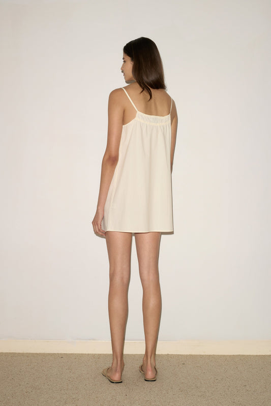 A woman faces away from the camera, wearing the Light Dress - Butter White by 0 and sandals, in a minimalist room with a plain wall and carpeted floor.