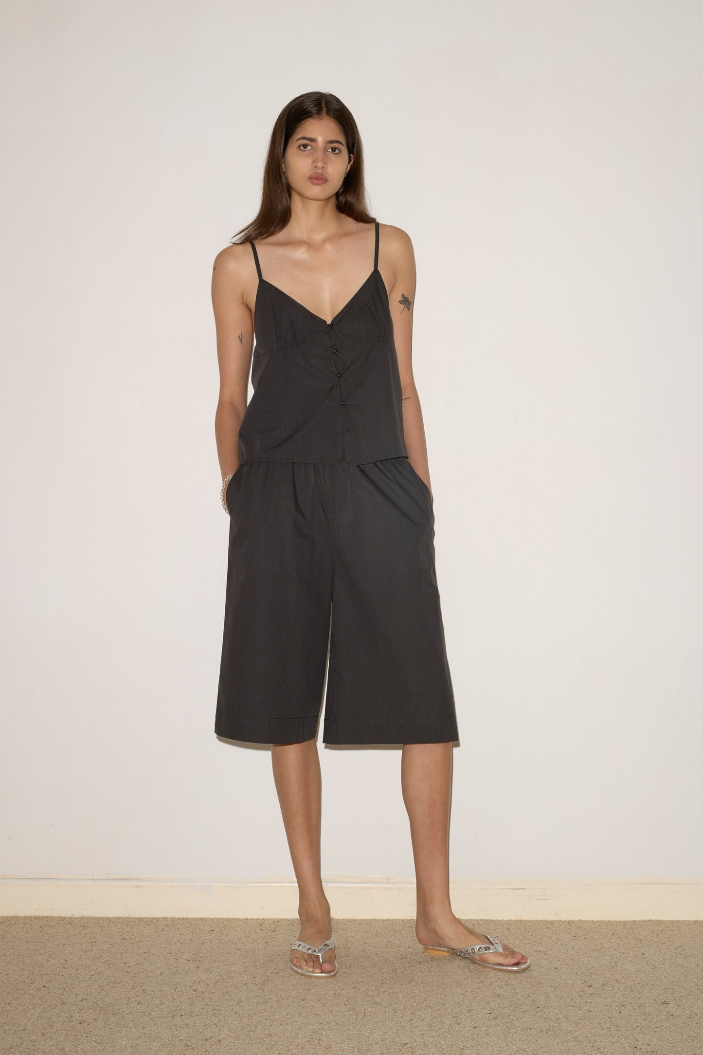 A woman with long hair stands against a plain light wall, wearing the 0 Seam Top - Black Stripe and matching black culotte shorts. She has her hands in her pockets and is wearing flip-flops.