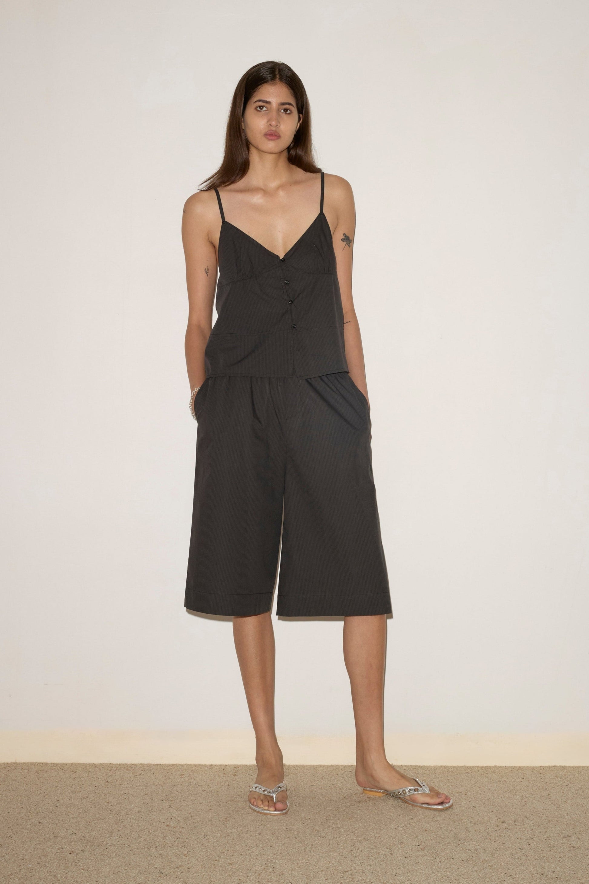A woman stands indoors against a plain wall, wearing Deiji Studios’ Long Lounger in Black Stripe—loose, sleeveless with thin straps and matching wide-leg shorts made from organic cotton. She has long hair, a neutral expression, and flat sandals.