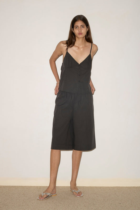 A woman with long brown hair stands against a plain wall, wearing the Long Lounger—a loose black sleeveless top with buttons and matching knee-length shorts. She pairs the outfit with flat sandals, her hands behind her back and looking forward.