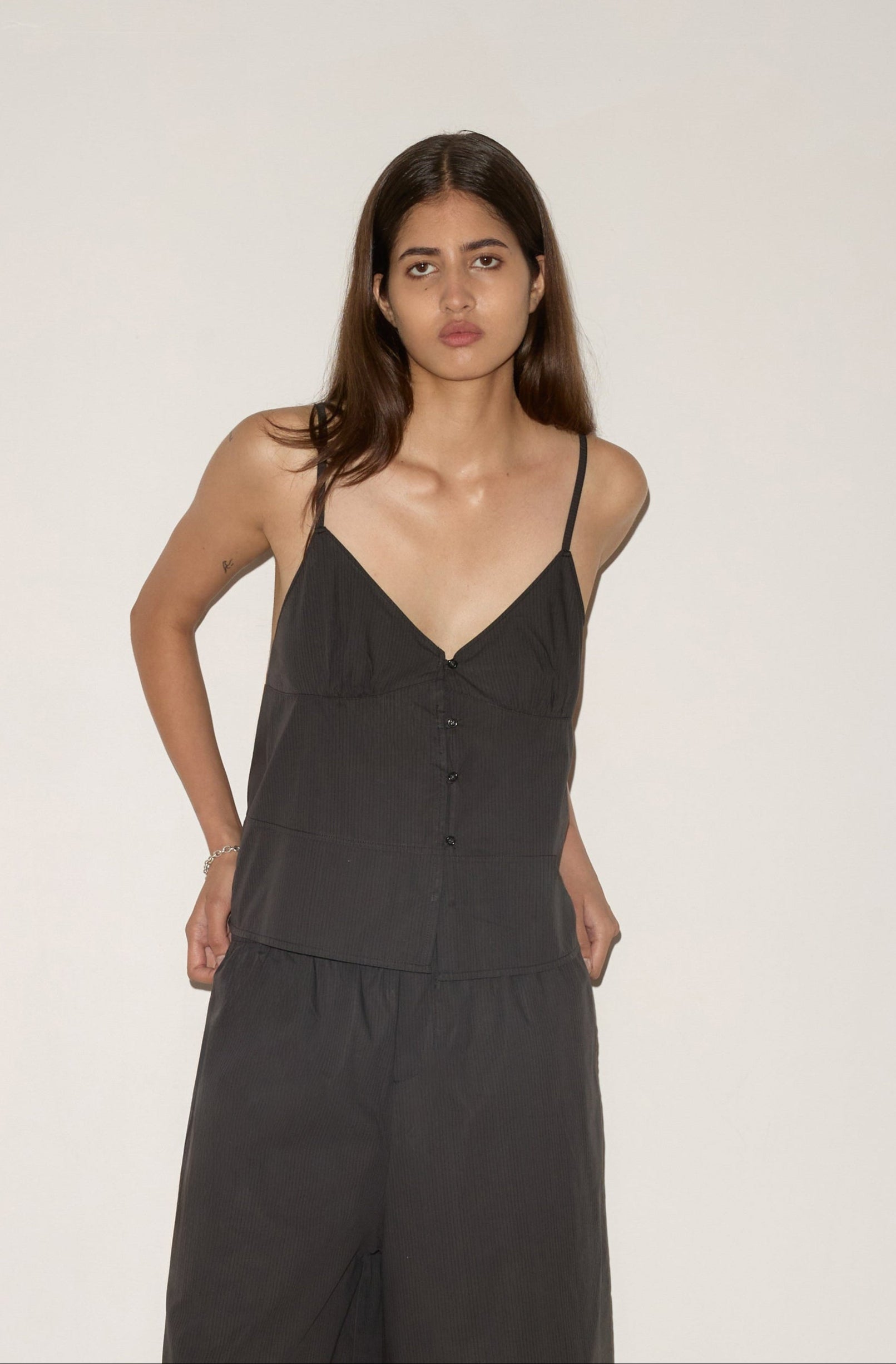 A woman with long brown hair stands against a plain light background, wearing the Seam Top—a black sleeveless top with thin straps—and matching loose black pants. She has a neutral expression and her hands are by her sides.