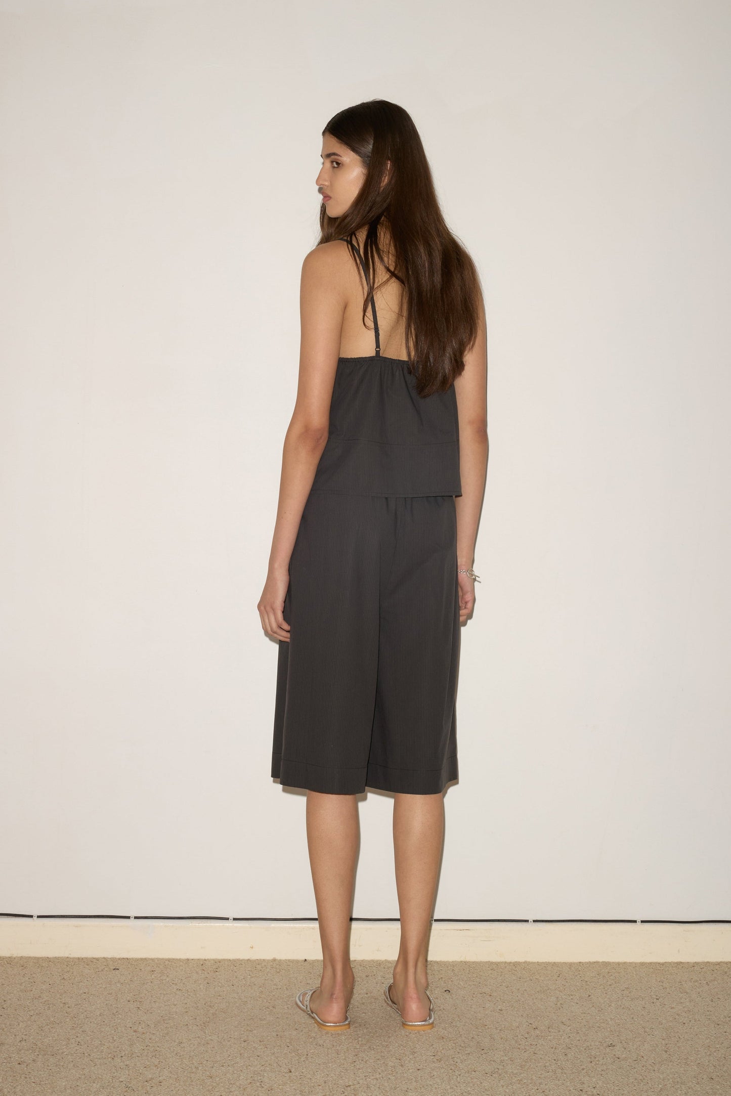A woman with long brown hair stands indoors, facing away from the camera in front of a light wall, wearing the 0 Seam Top - Black Stripe and matching knee-length shorts.