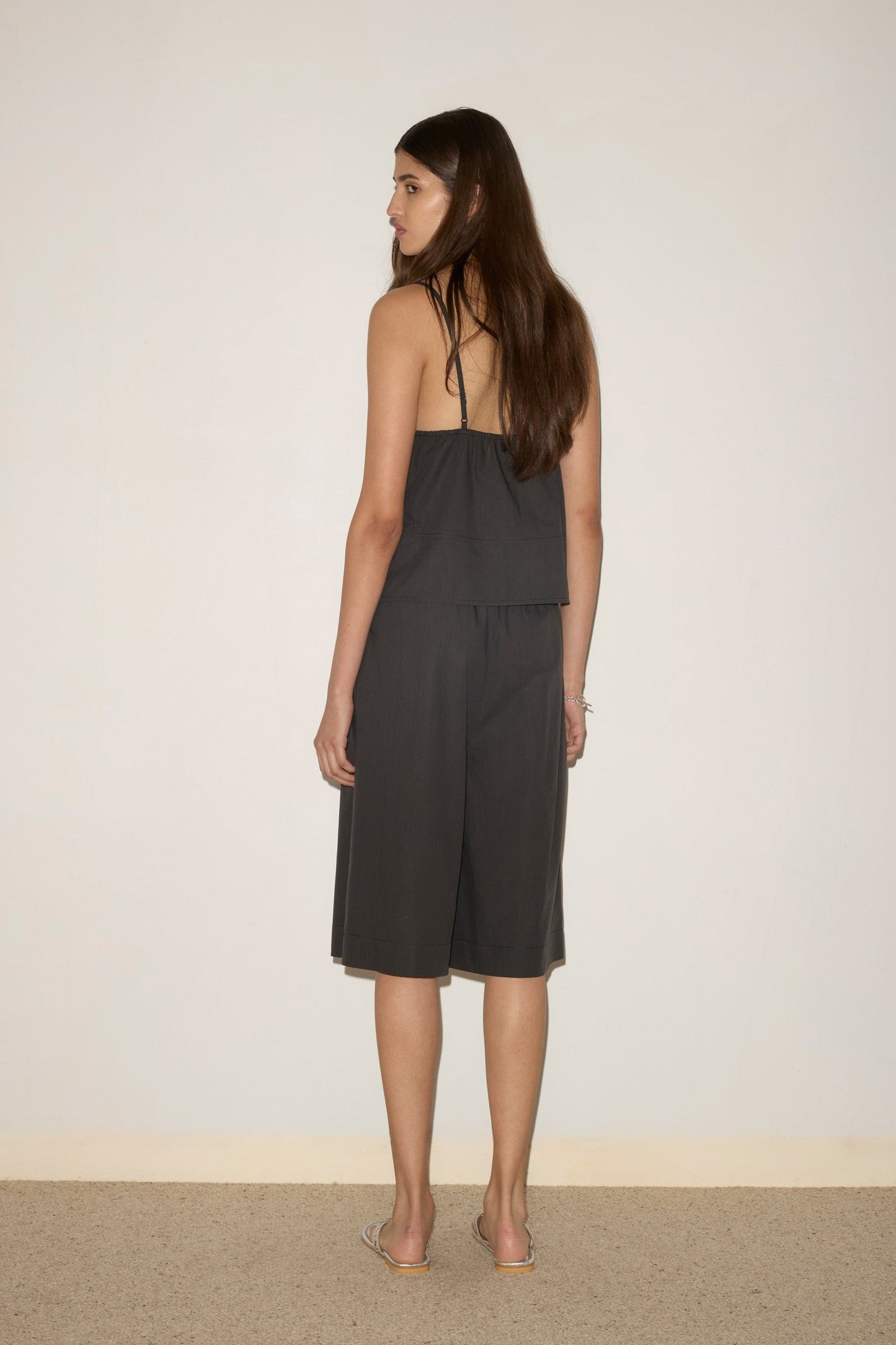 A woman with long brown hair stands facing away, wearing the Long Lounger - Black Stripe, a dark sleeveless knee-length outfit crafted from organic cotton, paired with silver sandals against a light wall and beige carpet.