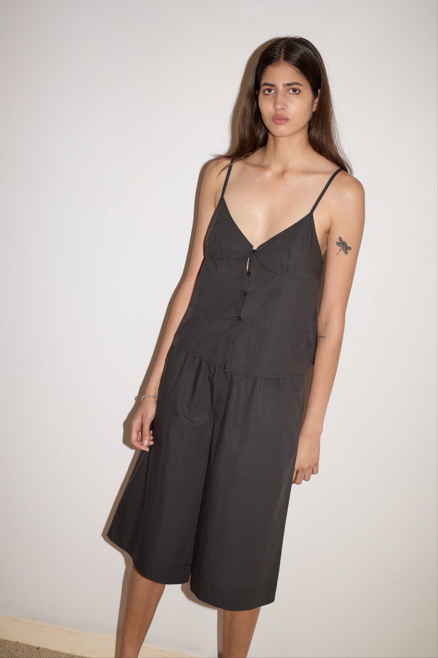 A woman with long brown hair wears the Seam Top - Black Stripe with adjustable straps and matching loose black culottes, standing against a plain light-colored wall. She has a bird tattoo on her upper left arm.
