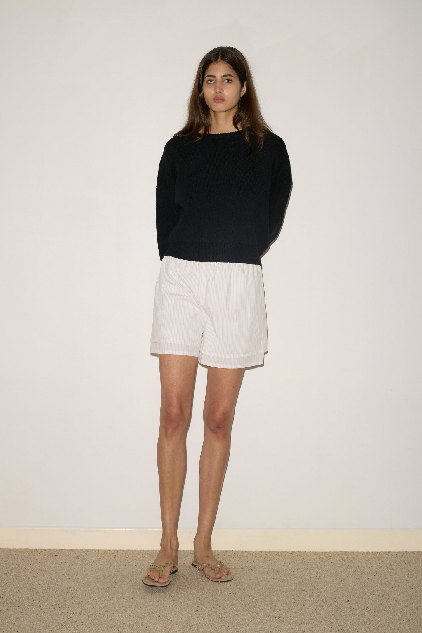 A woman with long brown hair stands against a white wall, wearing a black long-sleeve top, 0 Layered Short - Pacific Stripe, and flip-flops. Her hands are behind her back and she has a neutral expression.