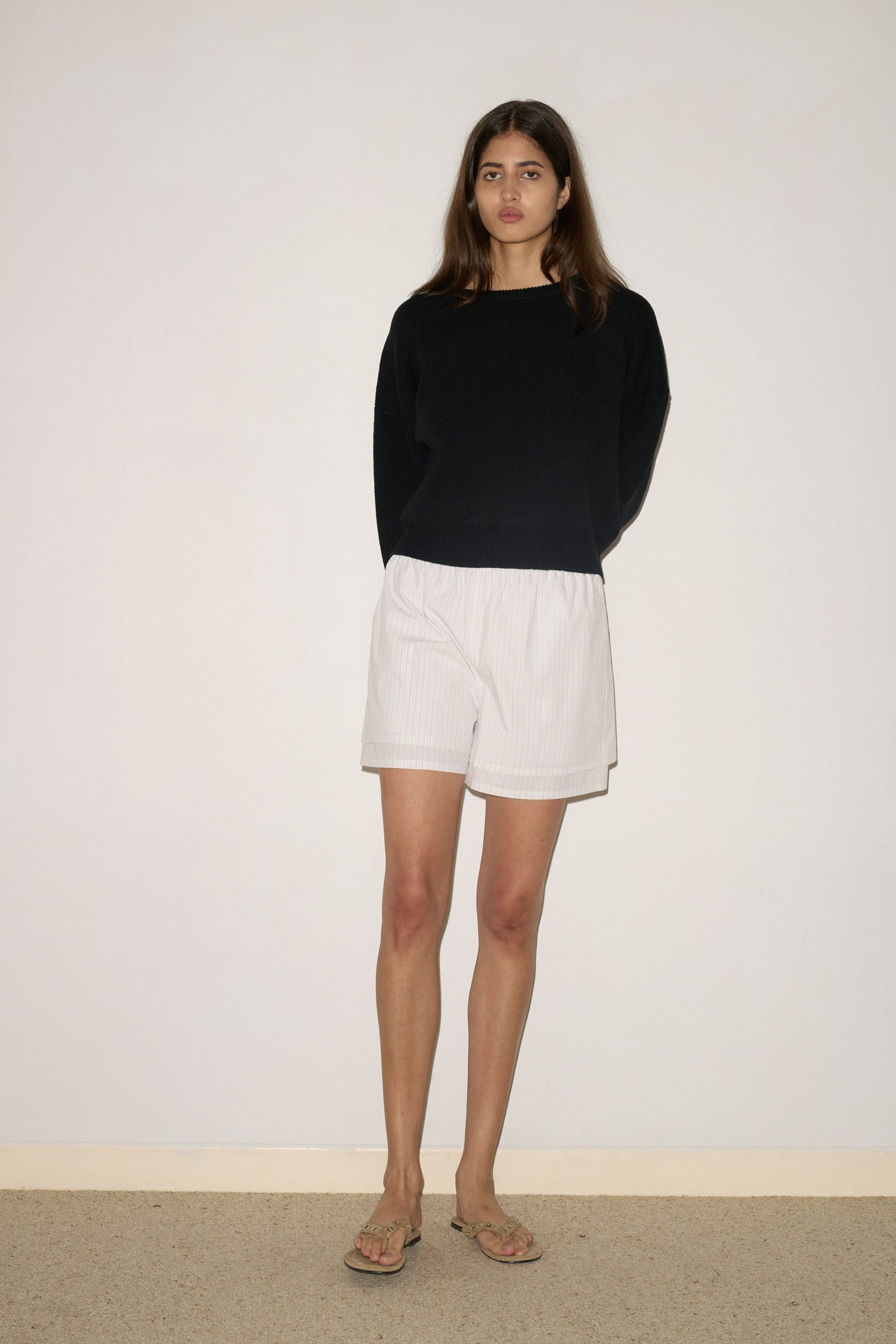 A woman with long brown hair stands against a light wall, wearing the Pullover - Black by 0, white shorts, and beige sandals. Her arms are behind her back and she maintains a neutral expression.