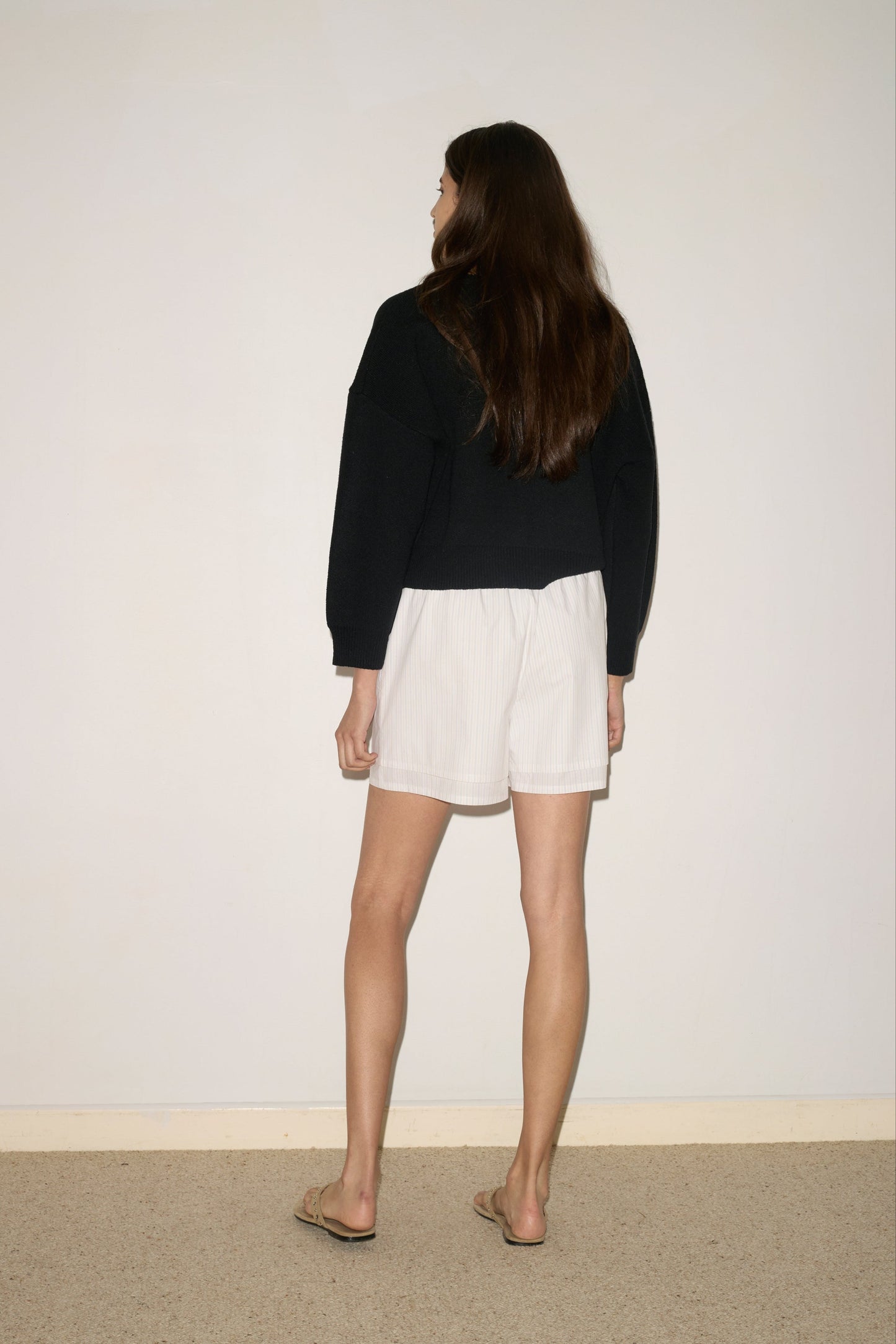 A woman with long brown hair stands facing a plain wall, wearing a black sweater, 0 Layered Short - Pacific Stripe, and flip-flops. She stands on a beige carpeted floor.
