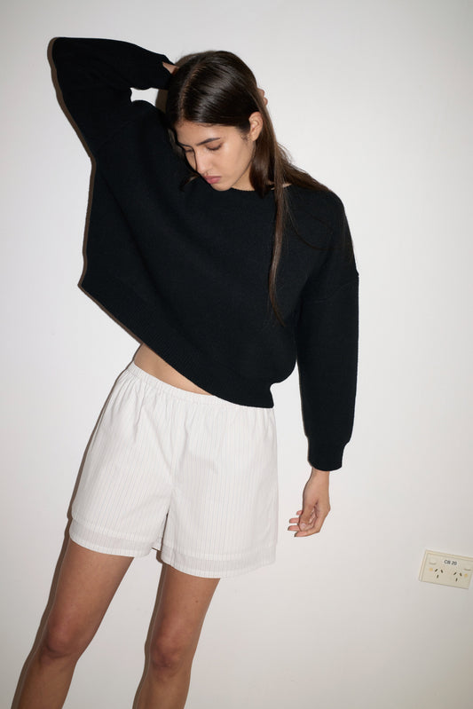 A young woman poses against a white wall wearing the Pullover, a loose black sweater paired with white shorts. She gazes downward, lifting one arm behind her head as her long hair tumbles over her shoulder.