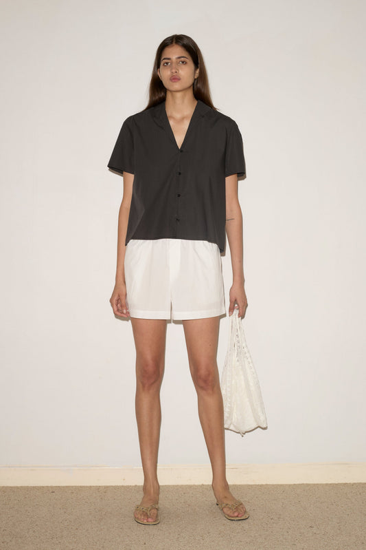 A woman stands against a plain wall, wearing the Sails Top - Black Stripe from Deiji Studios, white shorts, beige sandals, and holding a white textured bag.