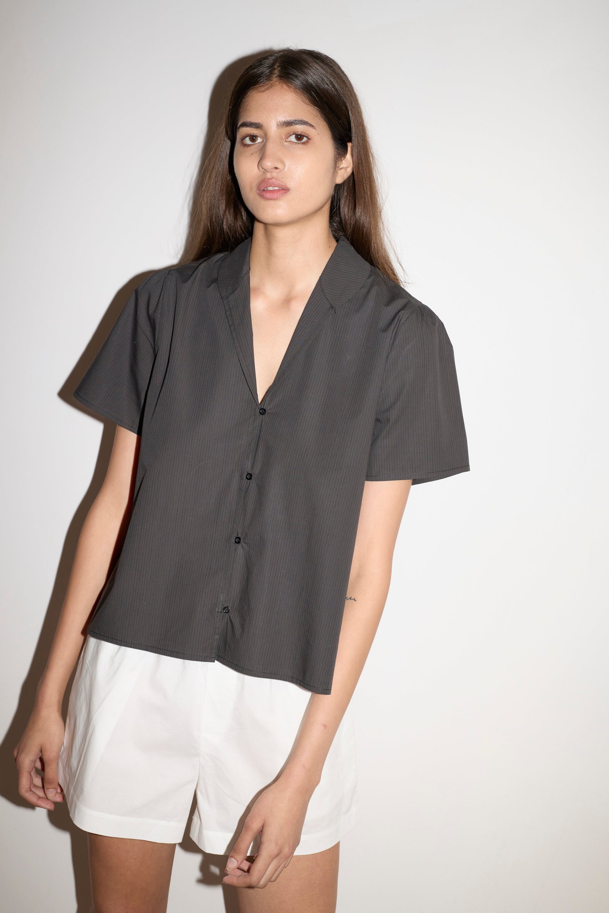 A woman with long brown hair wears the Sails Top - Black Stripe and white shorts, standing against a plain light background and looking calmly at the camera.
