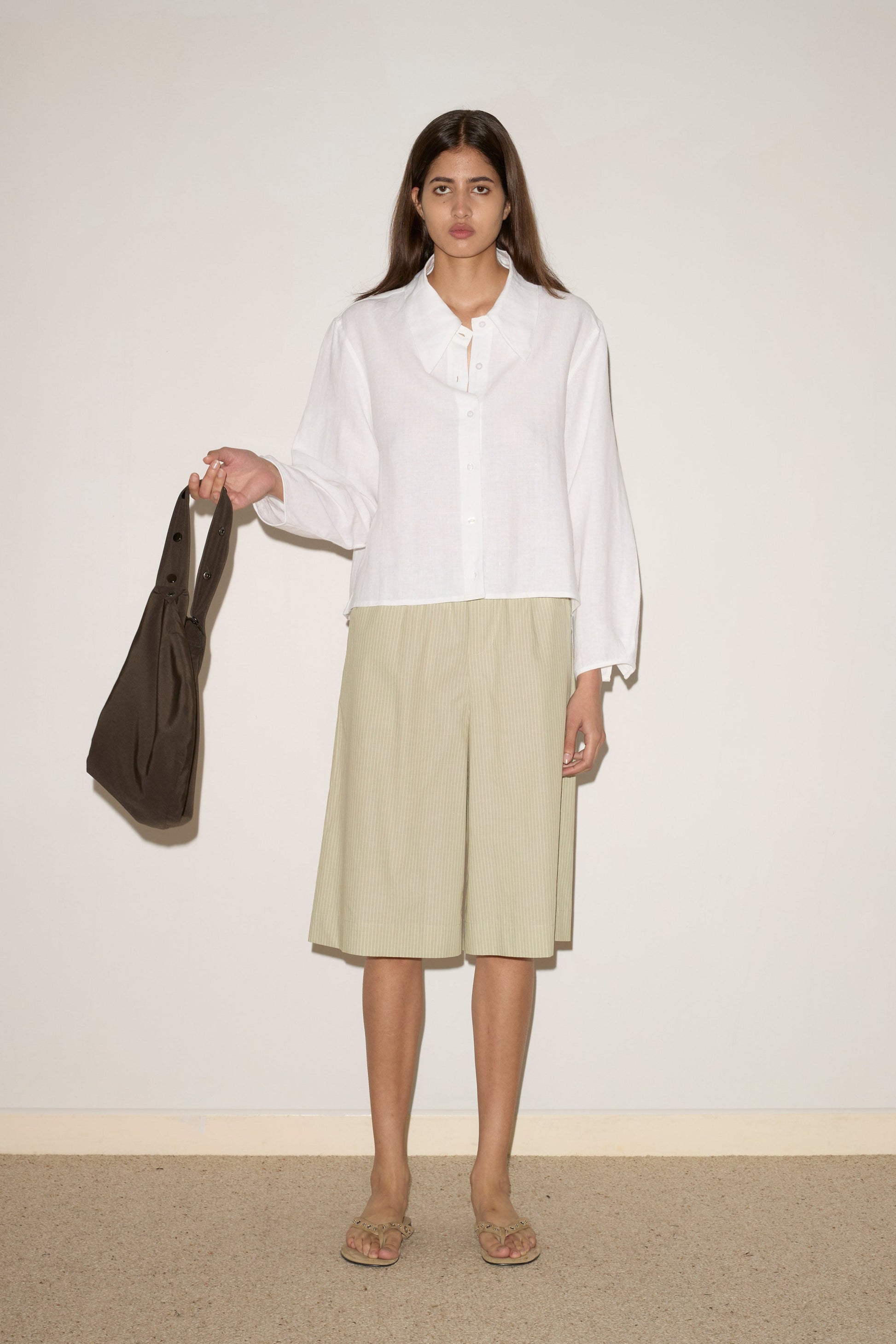 A woman stands against a plain wall wearing the Collar Point Shirt - White, light green knee-length shorts, and sandals. She holds a dark brown bag in her left hand.