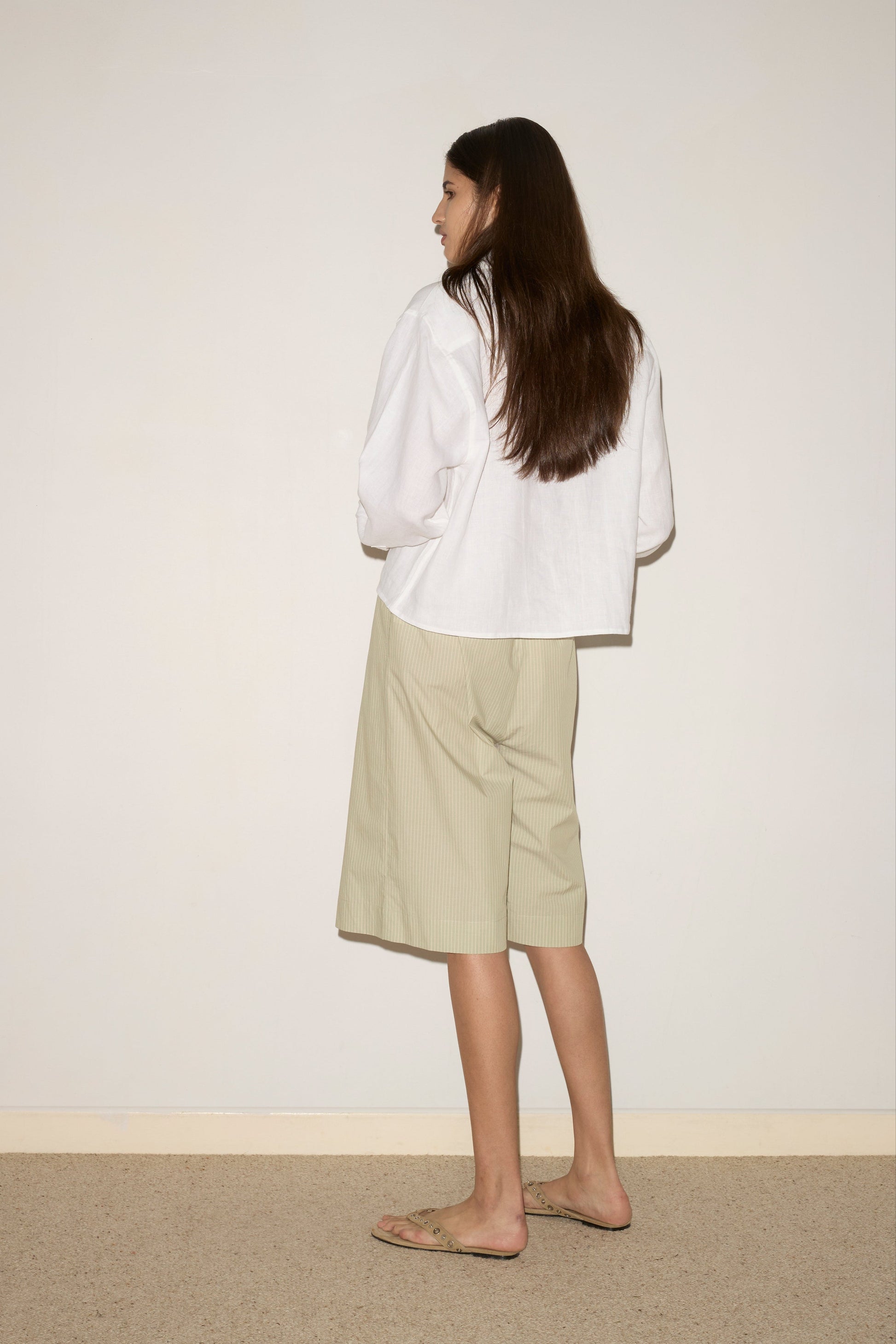 A woman with long brown hair, standing indoors on a neutral floor, wears the Collar Point Shirt - White, paired with light knee-length shorts and sandals as she faces a wall.