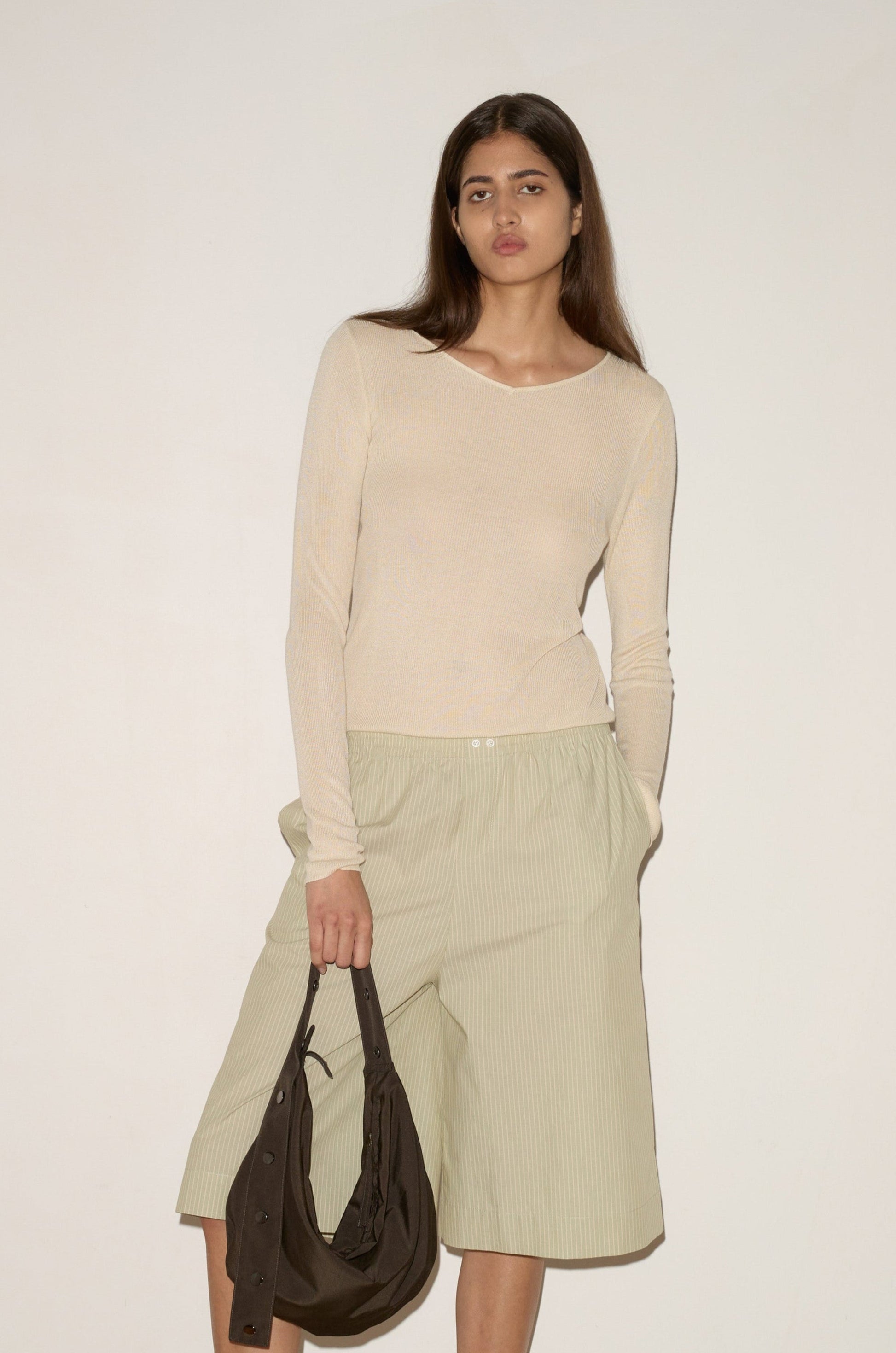 A woman with long brown hair wears the 0 Knitted Top - Fawn, featuring a fitted design and soft wide V neckline, paired with loose light green shorts. She stands against an off-white background holding a dark brown bag.