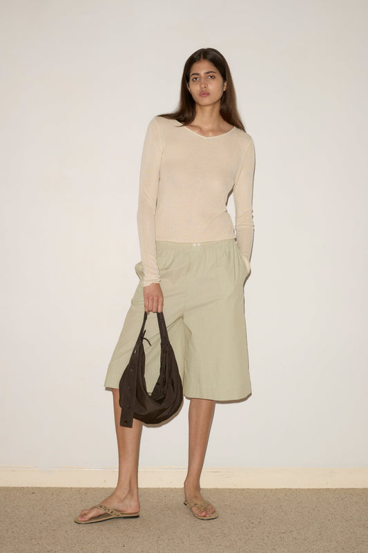 A woman with long dark hair and a neutral expression stands against a plain wall, wearing the Long Lounger - Pistachio Stripe, paired with beige sandals and holding a brown bag.