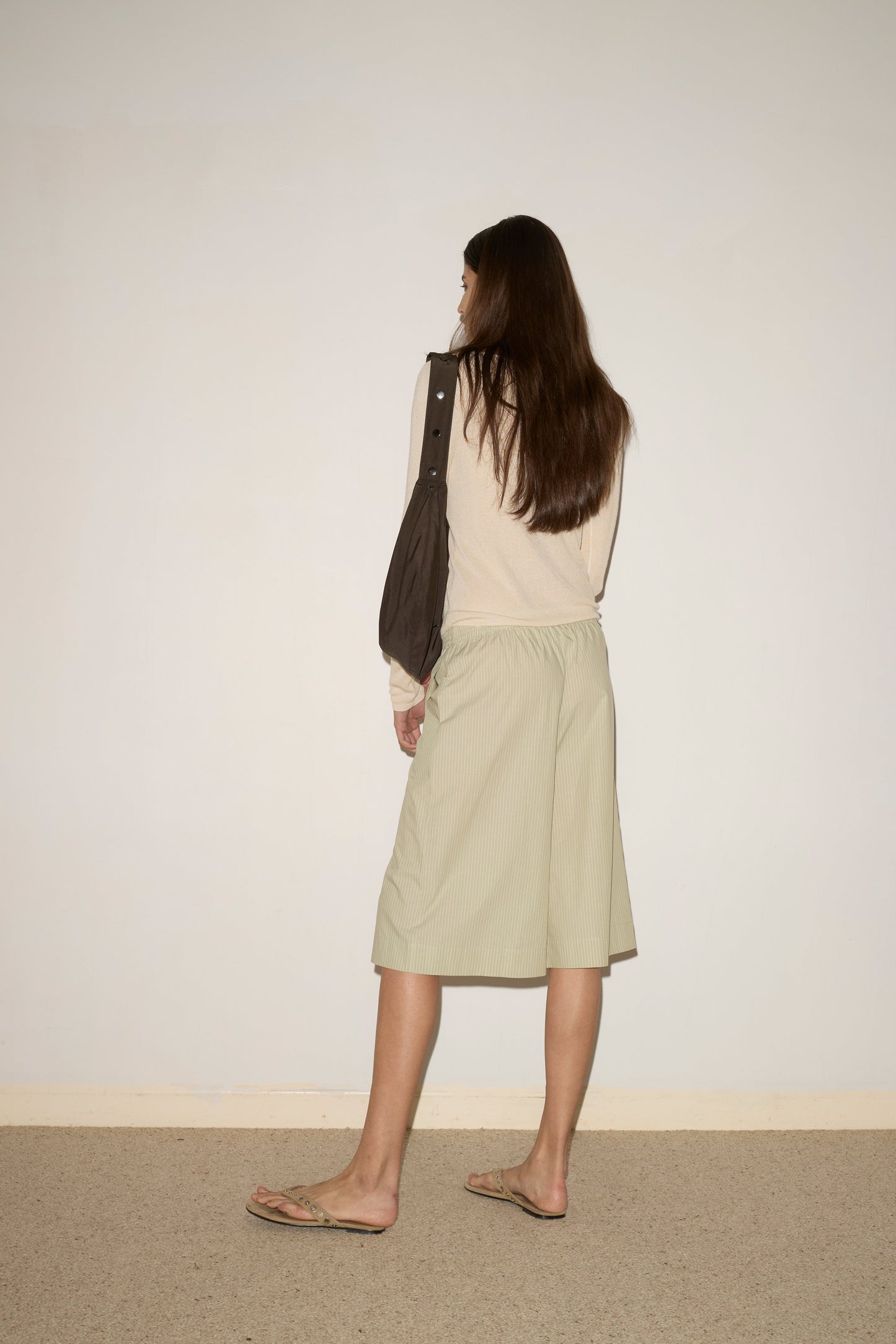A woman with long brown hair, wearing the Long Lounger - Pistachio Stripe by Deiji Studios, a dark shoulder bag, and flip-flops stands with her back to the camera, facing a blank wall.