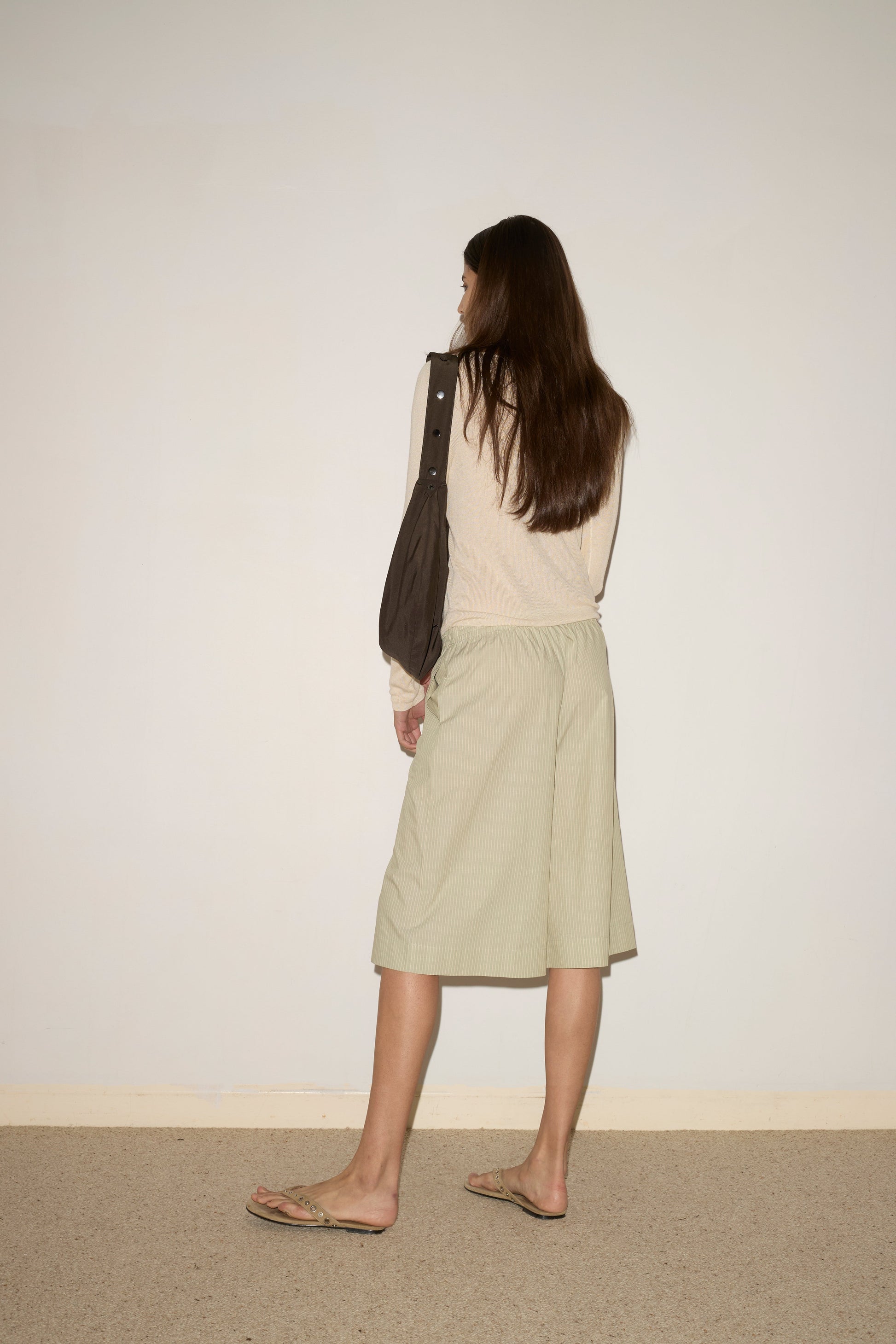 A woman with long brown hair, wearing the Long Lounger - Pistachio Stripe by Deiji Studios, a dark shoulder bag, and flip-flops stands with her back to the camera, facing a blank wall.