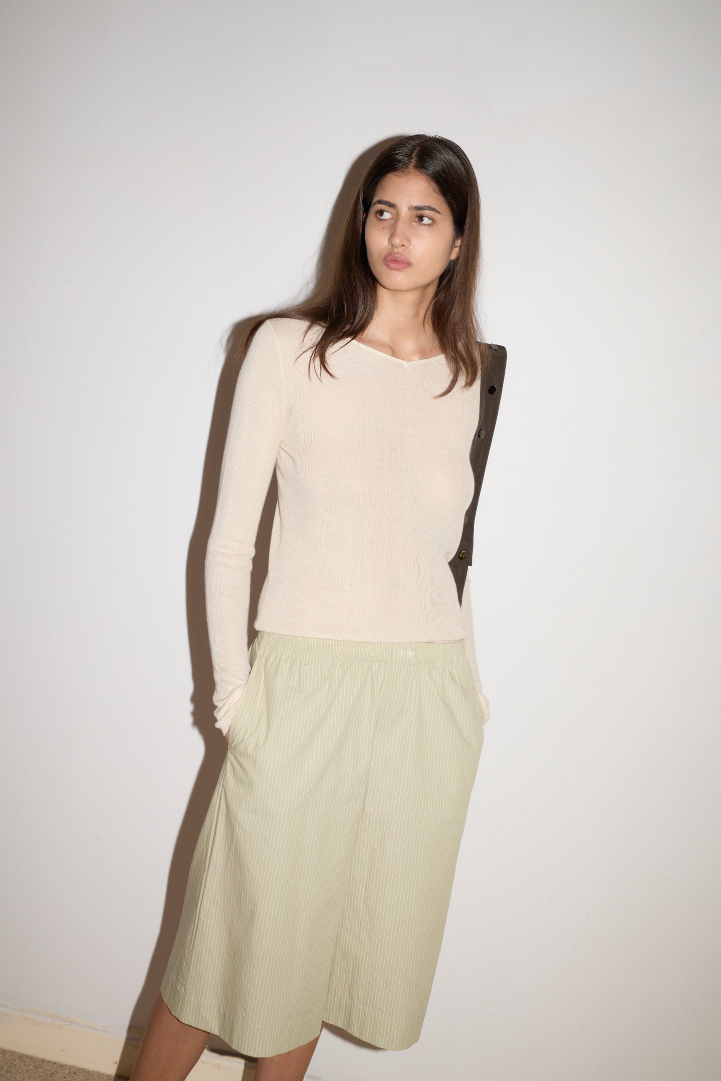A woman with long brown hair stands against a plain white wall, wearing the 0 Knitted Top - Fawn with a soft wide v neckline and loose pale green knee-length shorts, her hands in her pockets.
