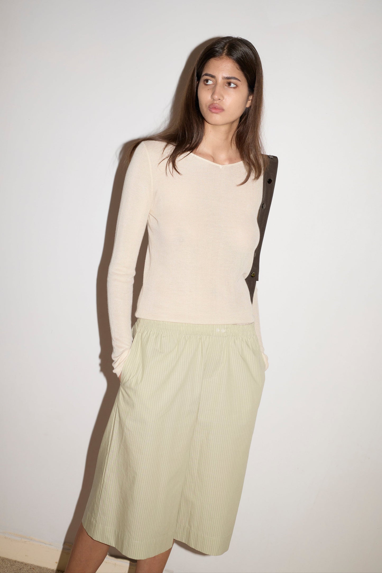 A woman with long brown hair stands against a white wall, wearing the Long Lounger—a beige long-sleeve top paired with a light green knee-length pleated skirt, hands in her pockets.