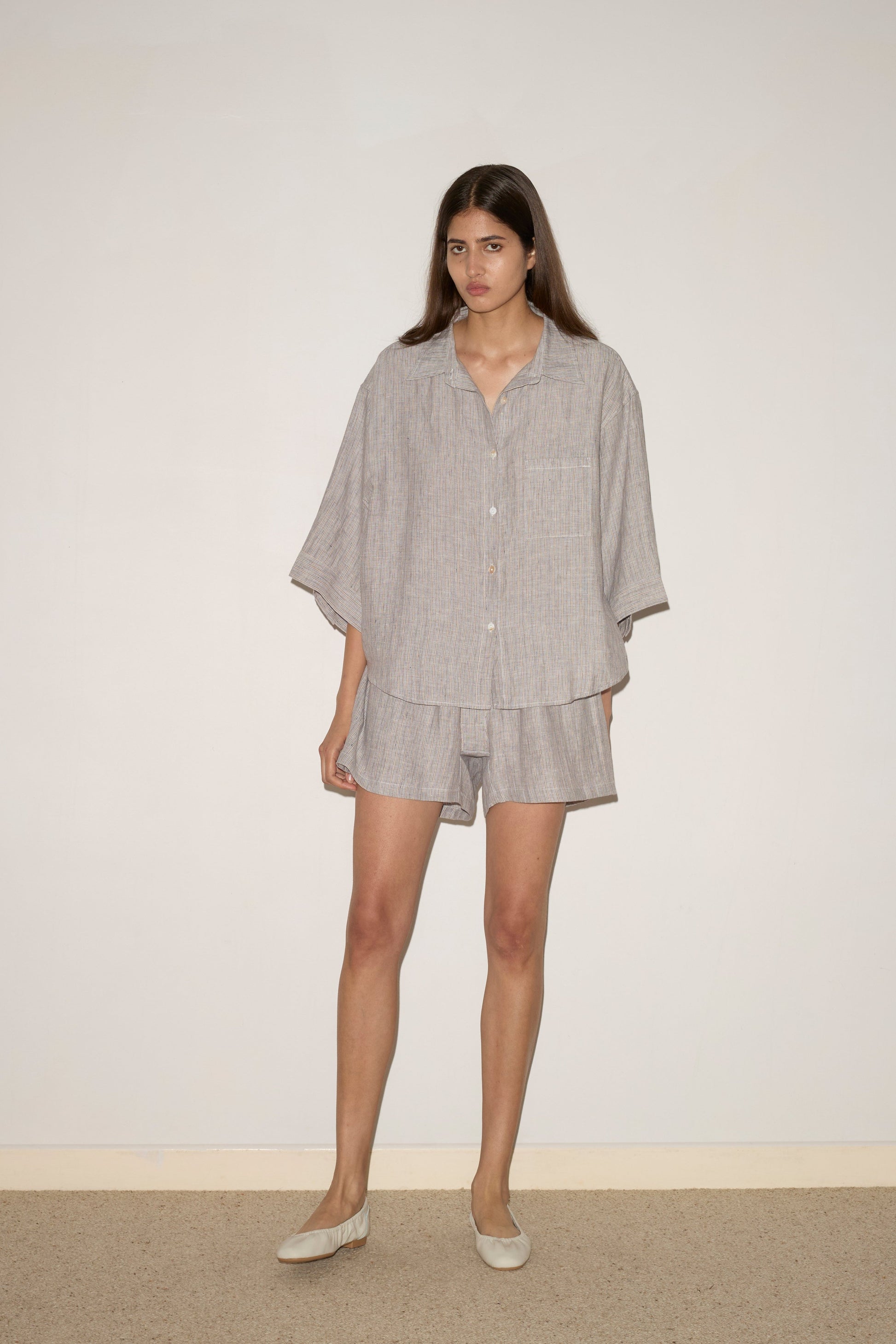 A woman with long brown hair stands against a plain wall wearing the 0 brands 03 Set - Haze Blue, featuring a loose GOTS certified linen shirt with three-quarter sleeves and matching shorts, paired with light-colored flat shoes.