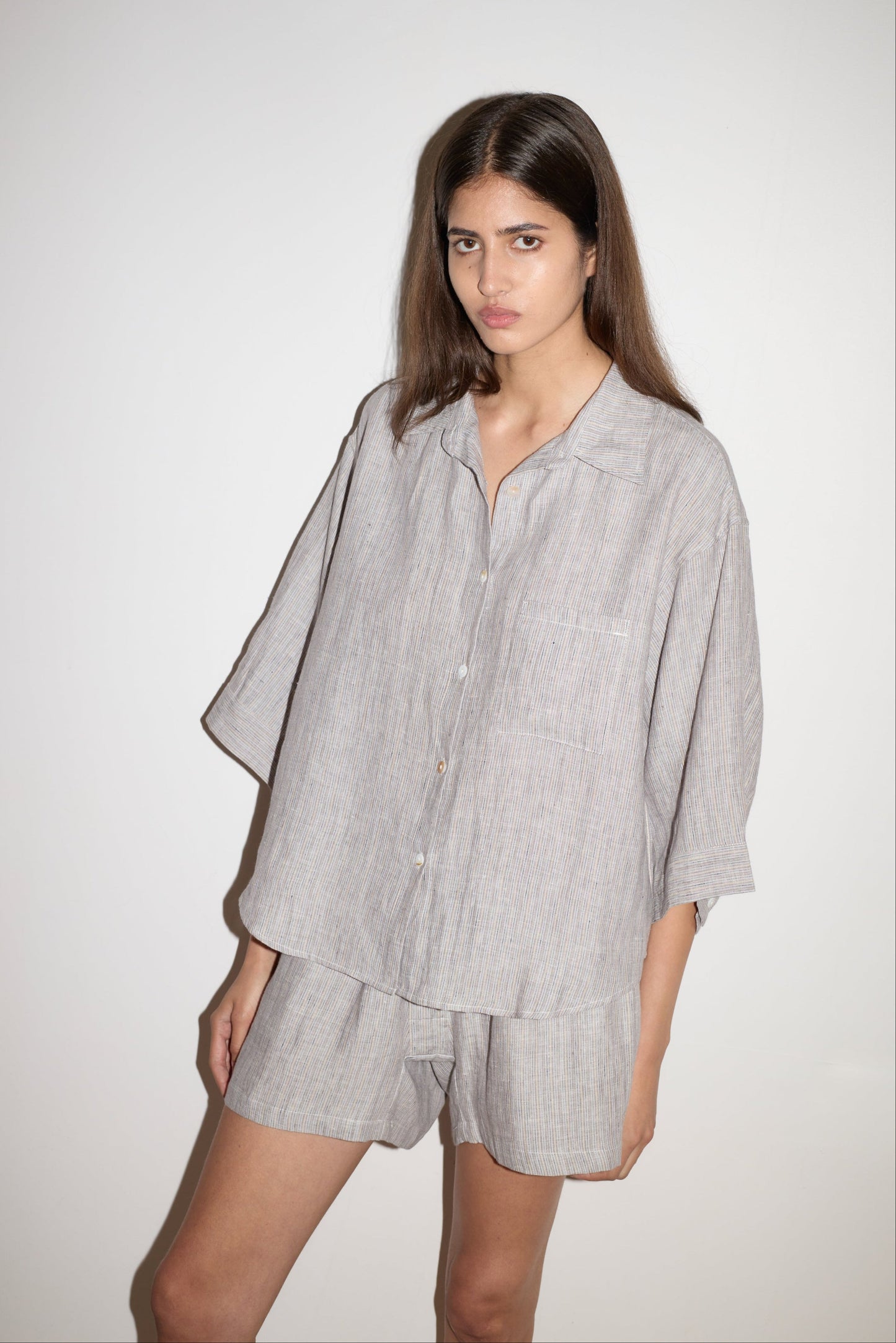 A woman with long brown hair wears the 03 Set - Haze Blue by 0, featuring a loose button-up shirt and matching shorts, while standing against a plain white background with a serious expression and her left hand resting on her thigh.