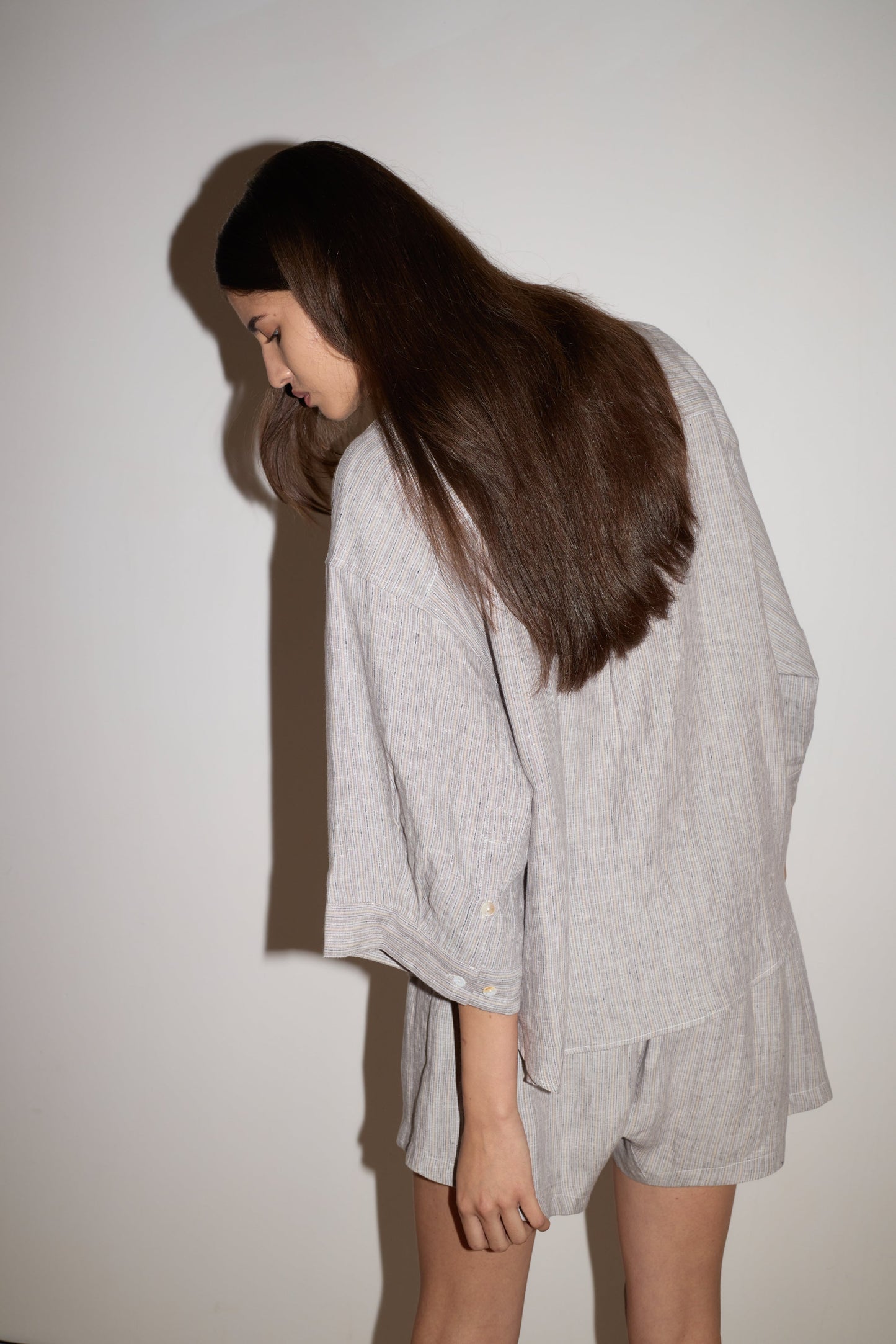 A woman with long, straight brown hair models the 0 brand’s 03 Set - Haze Blue, featuring a loose long-sleeve shirt and matching shorts, standing against a plain white wall and looking down with her body slightly turned away.
