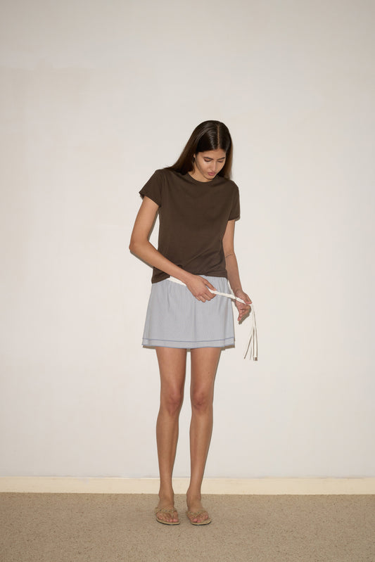 A woman with long brown hair wears the Layered Skirt - Sky Stripe, a brown organic cotton T-shirt, and sandals as she looks down to tie a white drawstring in front of a light-colored wall.
