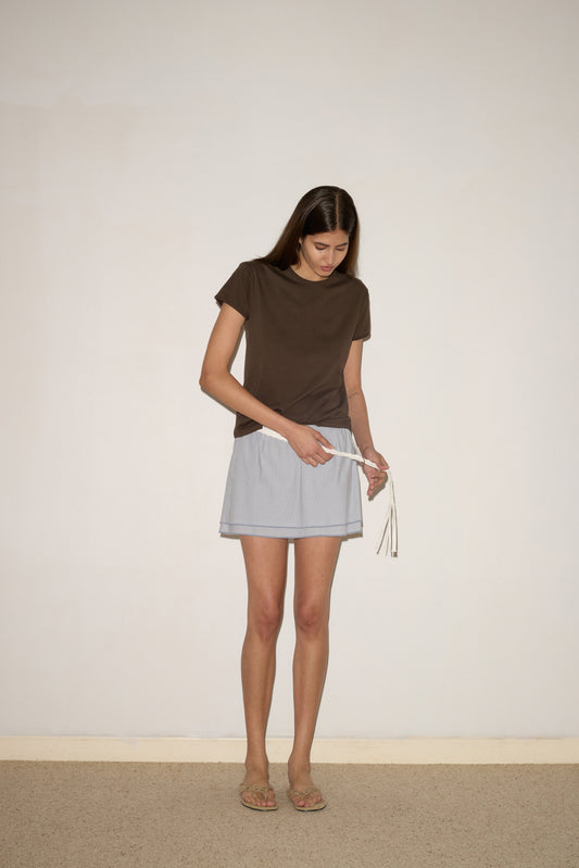 A woman with long brown hair stands indoors, looking down as she ties the white drawstring of her Deiji Studios Layered Skirt - Sky Stripe, paired with a brown t-shirt and sandals against a plain off-white wall.