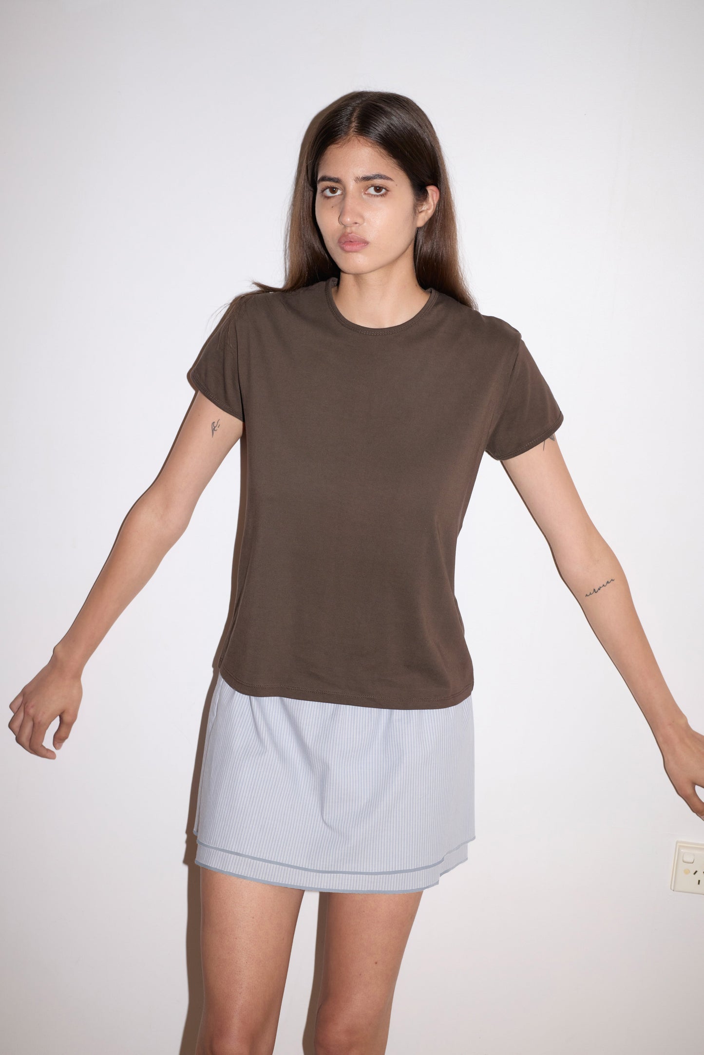 A woman with long brown hair stands against a white wall, wearing a brown t-shirt and the Layered Skirt - Sky Stripe from Deiji Studios. Her arms are slightly outstretched, and she has a neutral expression.