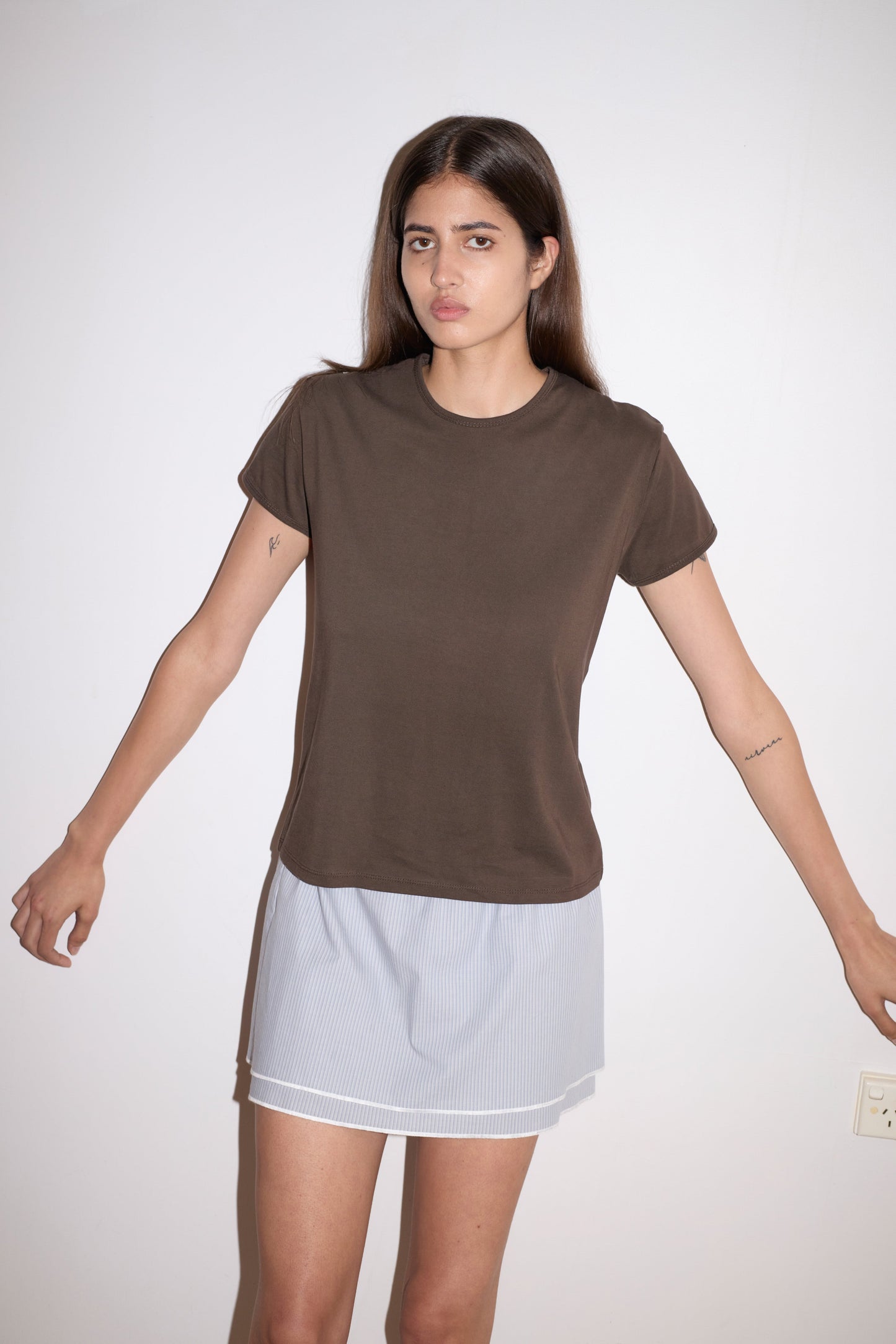 A woman with long brown hair stands against a plain white wall, wearing the Simple Tee in brown and a light skirt. Her arms are slightly extended and she has a neutral expression.