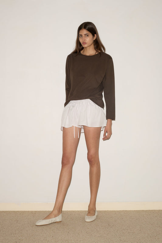 A woman stands indoors against a plain wall, wearing the Skort - Pacific Stripe by 0 and a long-sleeved brown top. She gazes slightly to the side with a neutral expression.