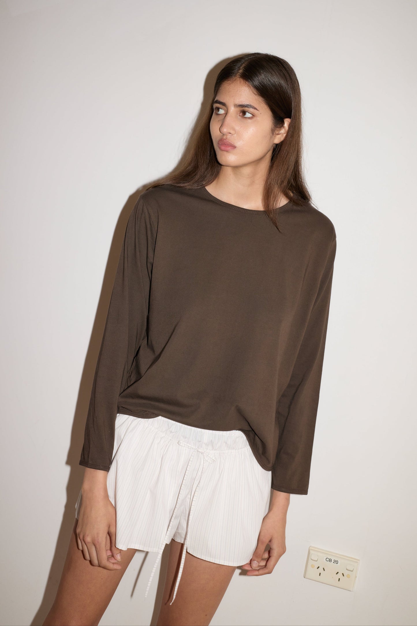 A young person with long, straight brown hair wears the 0 Simple Long Sleeve - Caribou and white shorts, standing against a plain white wall with a power outlet visible near their hand.