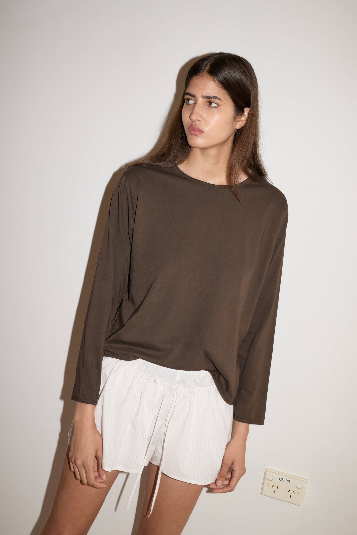 A woman with long brown hair stands against a white wall, wearing a loose brown long-sleeve top and the 0 Skort - Pacific Stripe. She gazes slightly to the side with a neutral expression.