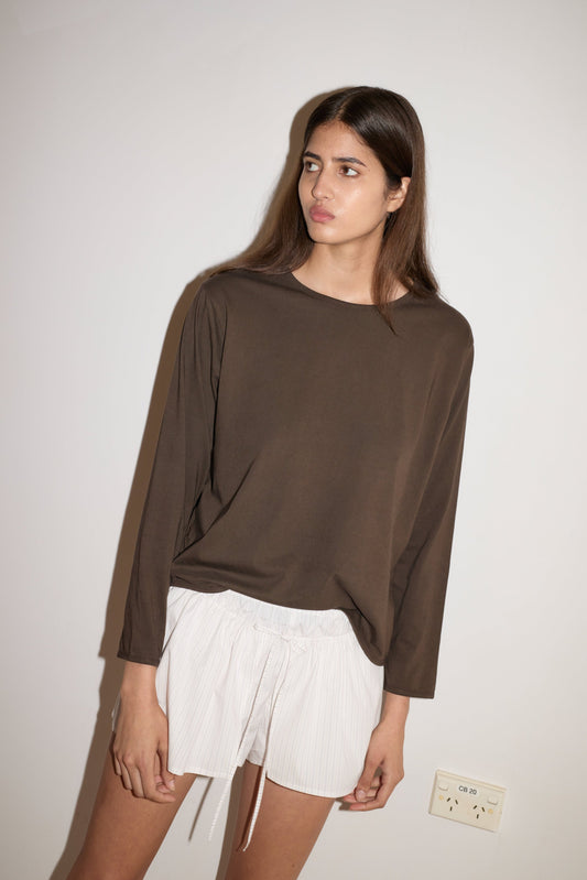 A woman with long brown hair stands against a white wall, wearing a loose brown long-sleeve top and the 0 Skort - Pacific Stripe. She gazes slightly to the side with a neutral expression.