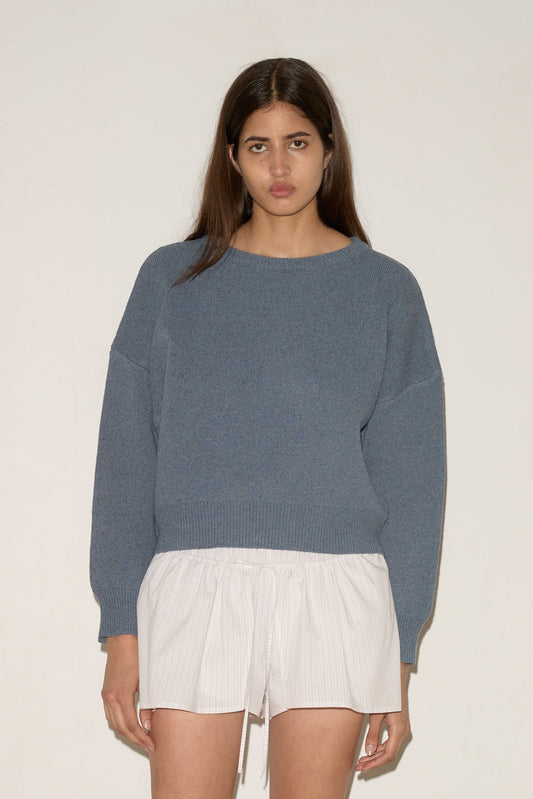 A woman with long brown hair and a neutral expression models the 0 Pullover - Glacier Blue against a white wall, styled with white drawstring shorts and light-colored flats.