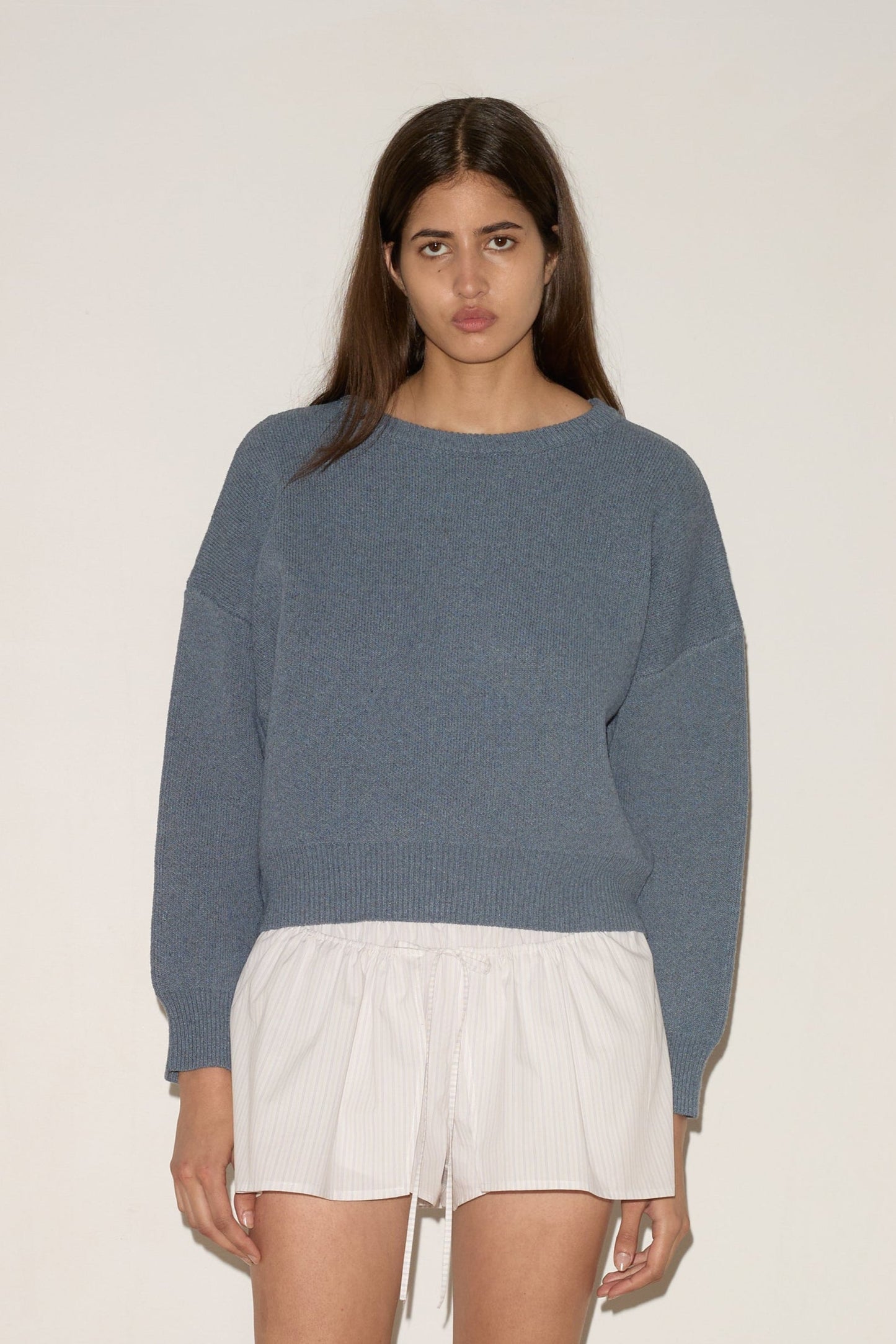 A woman with long brown hair and a neutral expression models the 0 Pullover - Glacier Blue against a white wall, styled with white drawstring shorts and light-colored flats.