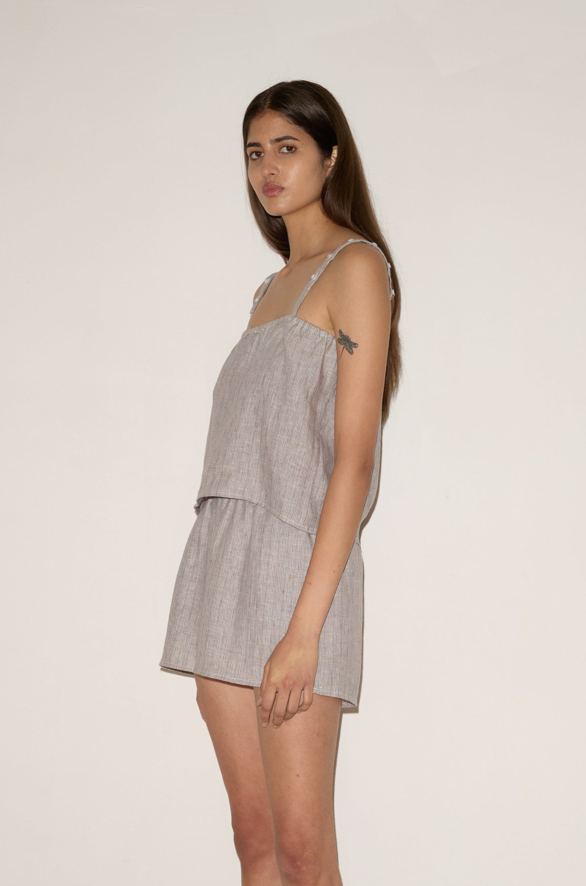 A young woman with long brown hair stands sideways against a light wall, wearing the 0 Button Linen Set in Haze Blue and sandals. She looks calmly at the camera, and beige carpet covers the floor.