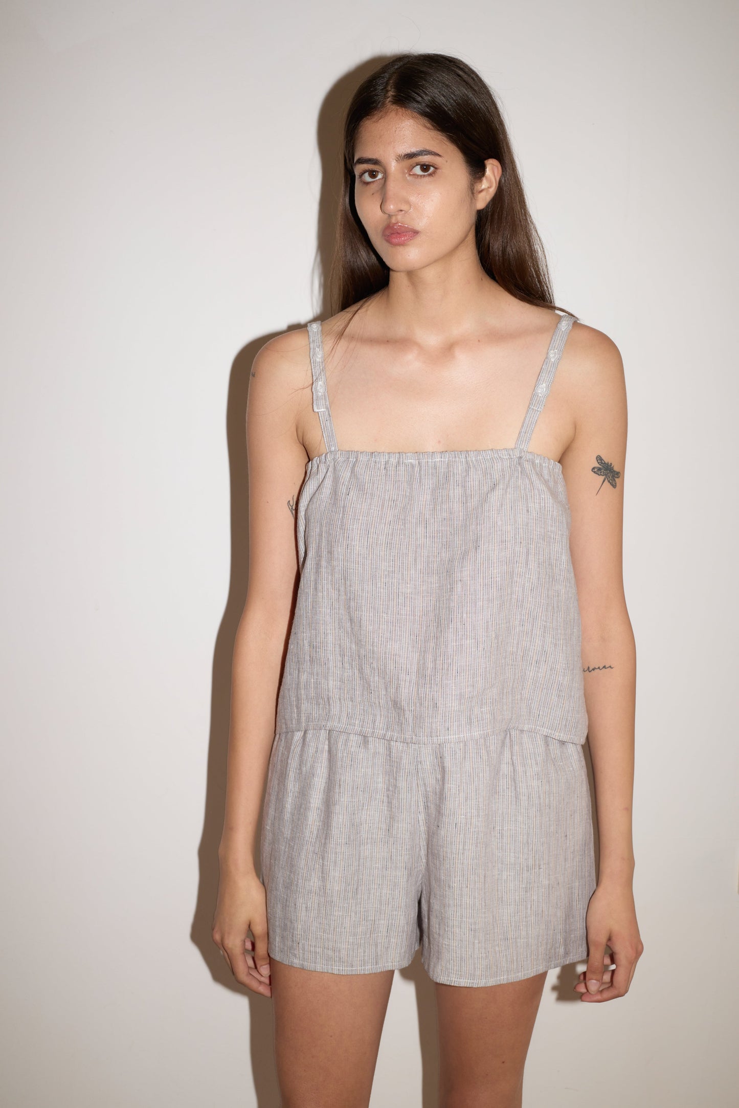 A woman with long brown hair and visible tattoos on her left arm wears the Button Linen Set - Haze Blue by 0, standing against a white wall and looking at the camera with a neutral expression.