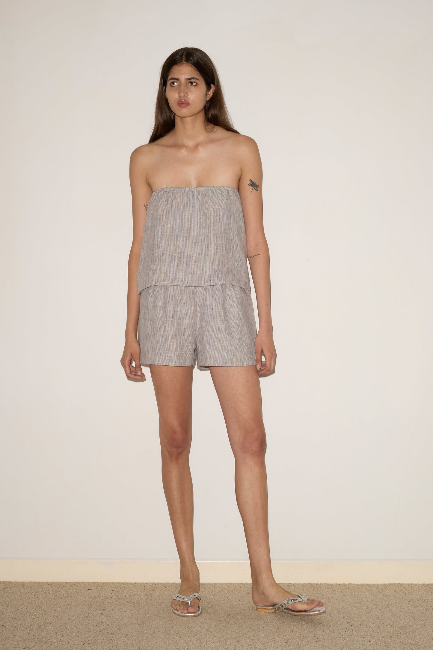 A woman with long brown hair stands against a plain wall, wearing the 0 Button Linen Set in Haze Blue. She has a tattoo on her upper left arm, flip-flops, and a neutral expression.