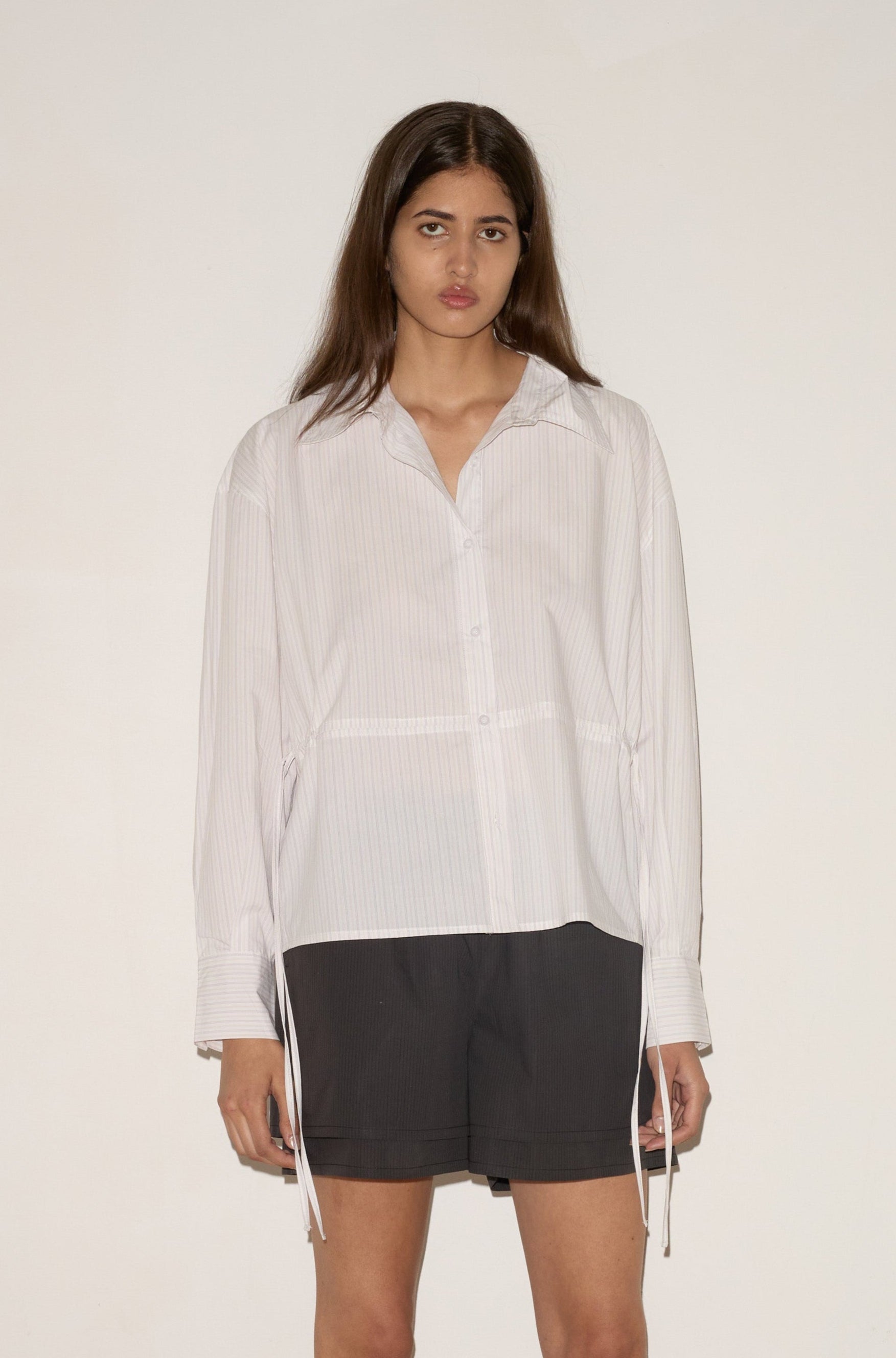 A woman with long brown hair stands against a white wall, wearing a loose button-up shirt and the Layered Short - Black Stripe by 0. She looks straight ahead with a neutral expression.