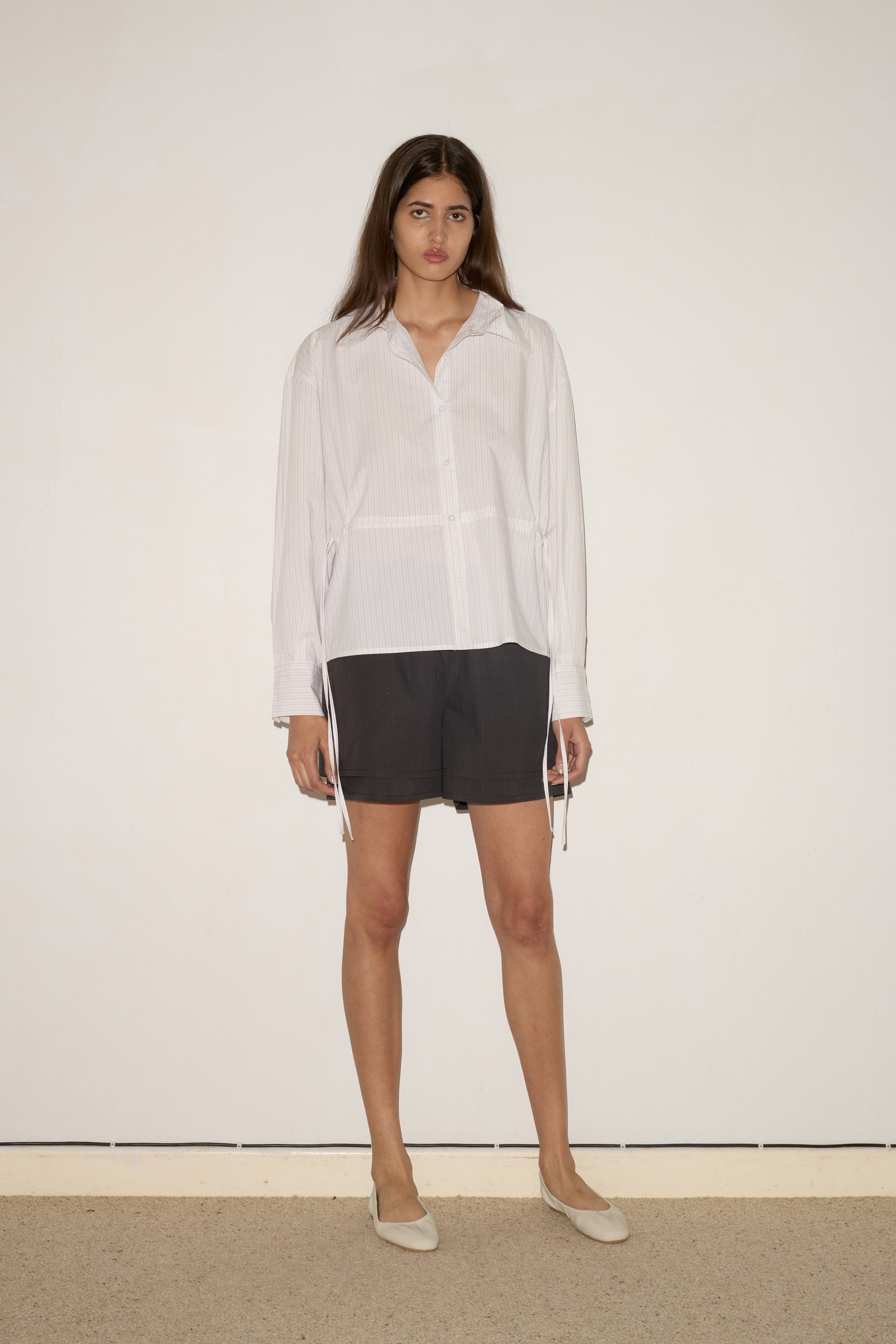 A woman with long brown hair stands against a white wall, wearing the 0 Room Shirt - Pacific Stripe with long sleeves, dark knee-length shorts, and light flats. She faces forward with a neutral expression.