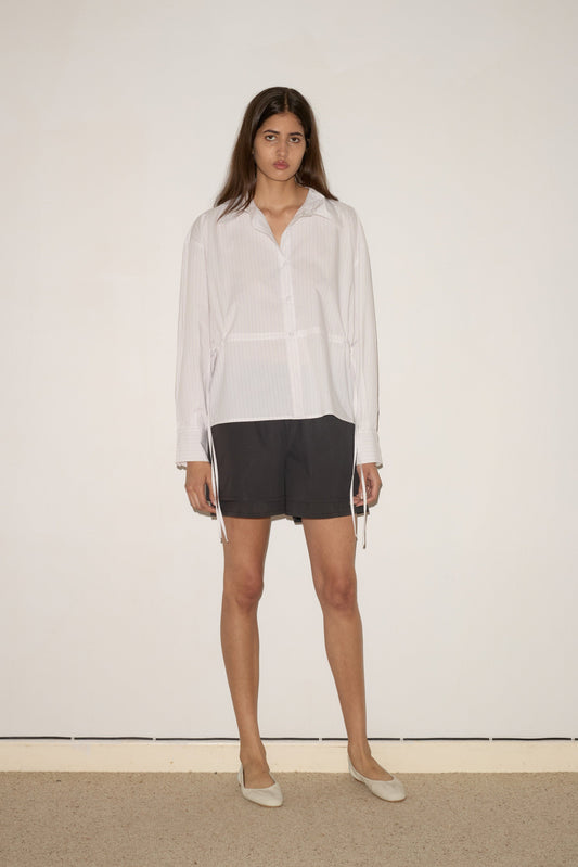 A woman with long brown hair stands against a white wall, wearing the 0 Room Shirt - Pacific Stripe with long sleeves, dark knee-length shorts, and light flats. She faces forward with a neutral expression.