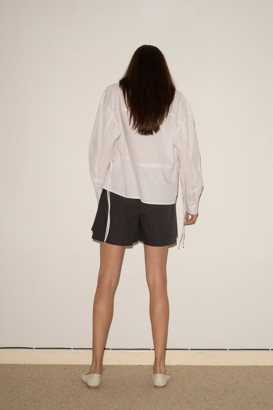 A person with long brown hair faces a plain white wall, wearing the Room Shirt - Pacific Stripe from 0, black shorts, and white slip-on shoes. Their arms rest relaxed at their sides.