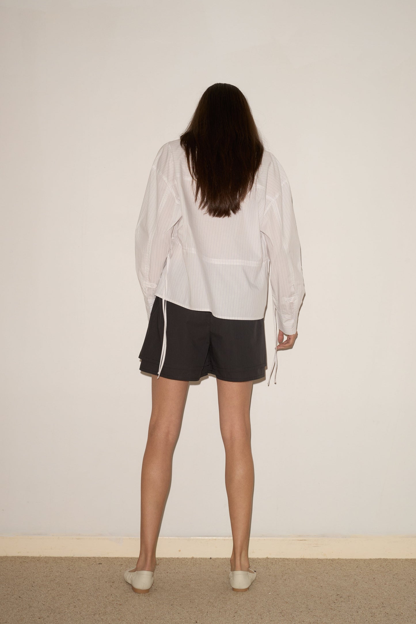 A person with long brown hair stands indoors, facing a plain light wall. They wear a white long-sleeve shirt, the Layered Short - Black Stripe with an elastic waist, and light slip-on shoes. Their back is visible in the photo.