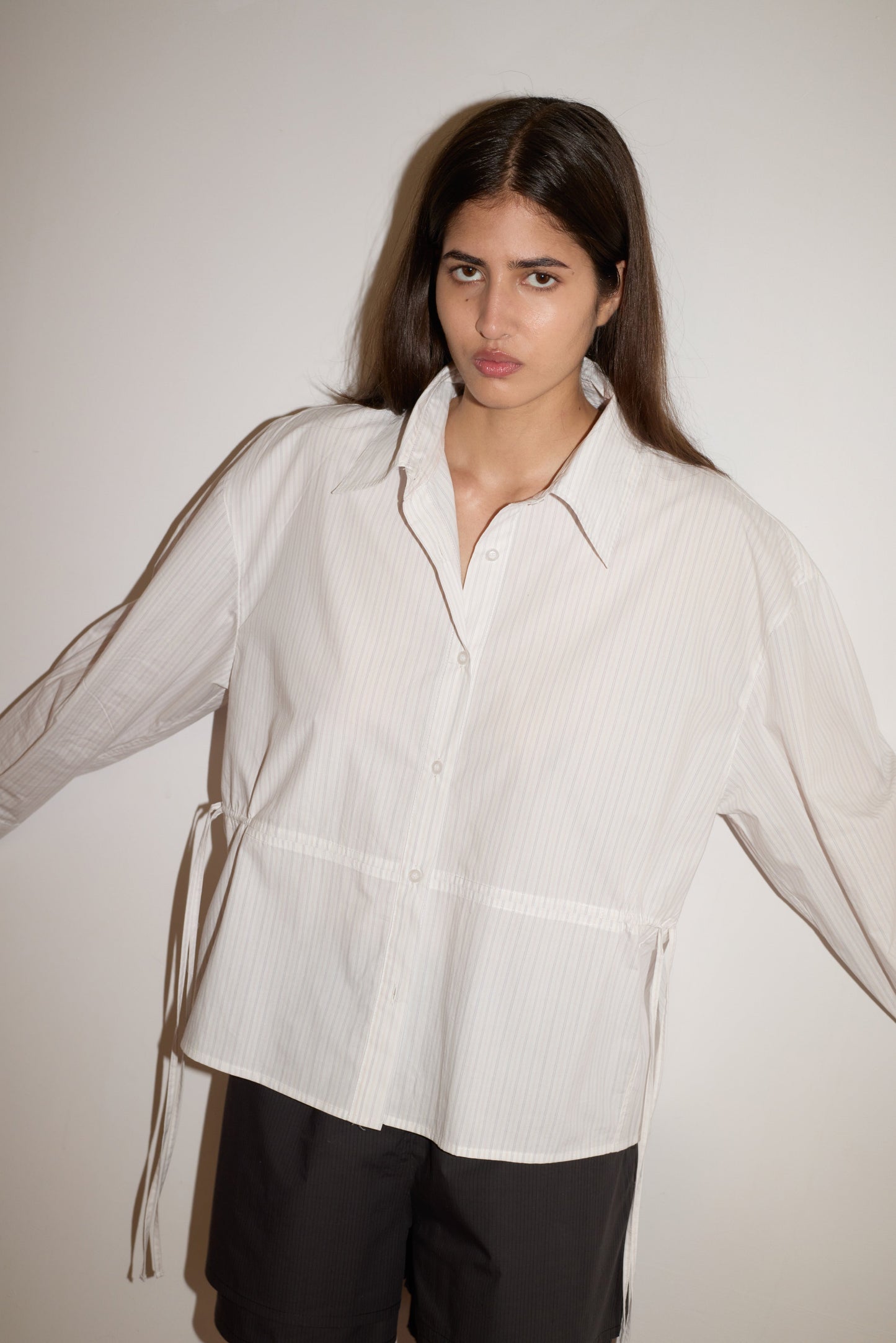 A woman with long brown hair stands against a white wall, wearing the oversized Room Shirt—light-colored with subtle stripes—and black shorts. Her arms are slightly outstretched and her expression is neutral.