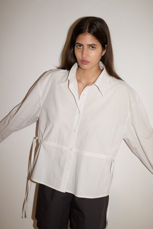 A woman with long brown hair stands against a white wall, wearing the oversized Room Shirt—light-colored with subtle stripes—and black shorts. Her arms are slightly outstretched and her expression is neutral.