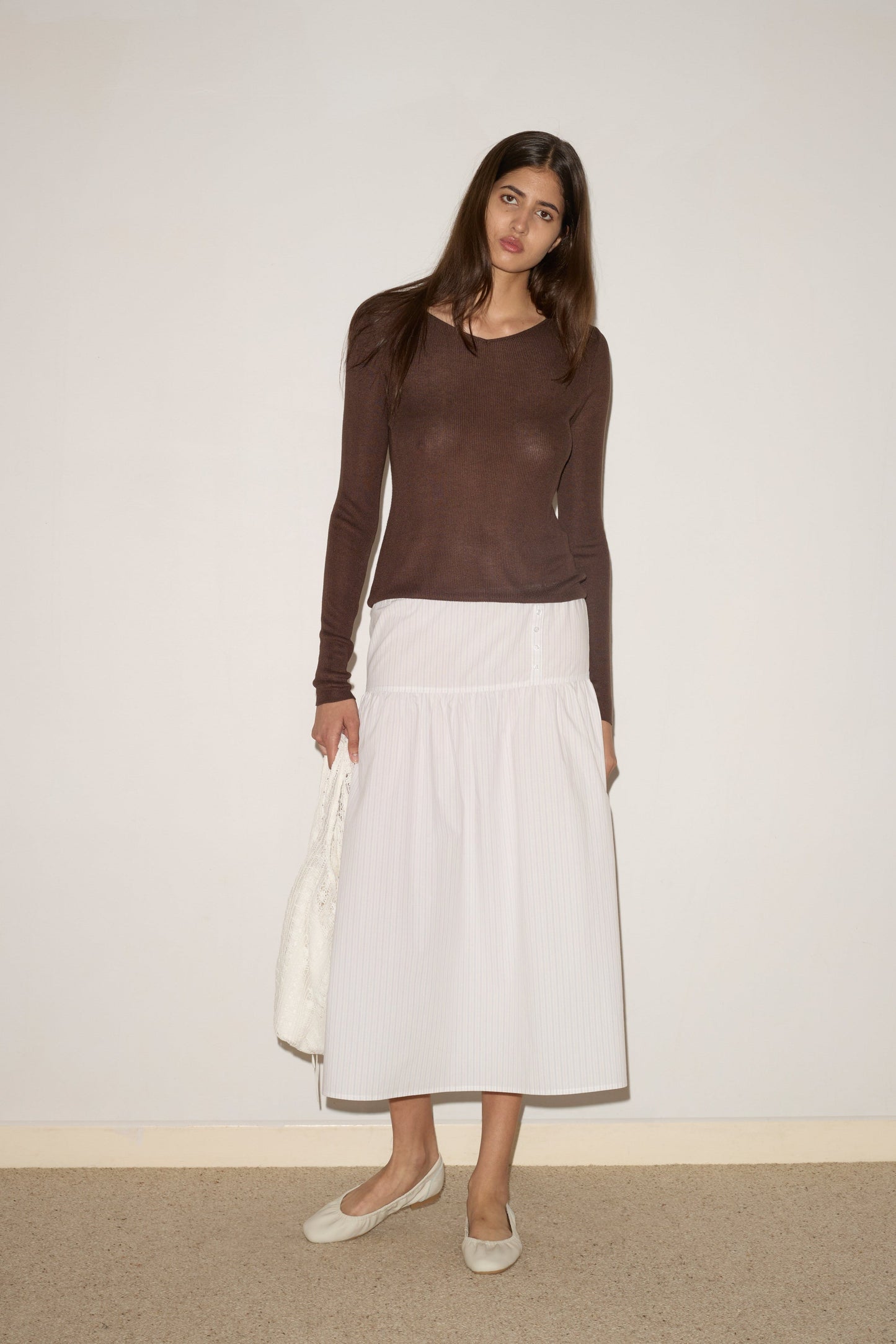 A woman with long brown hair stands against a plain wall, wearing the Button Skirt - Pacific Stripe by 0, paired with a sheer brown long-sleeve top, beige flats, and holding a white textured bag as she looks slightly to the side.