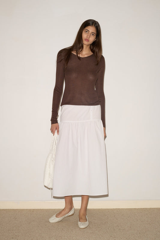 A woman with long brown hair stands by a plain wall, wearing 0’s Knitted Top in Dark Brown, a white ankle-length skirt, white flats, and holding a white textured bag.