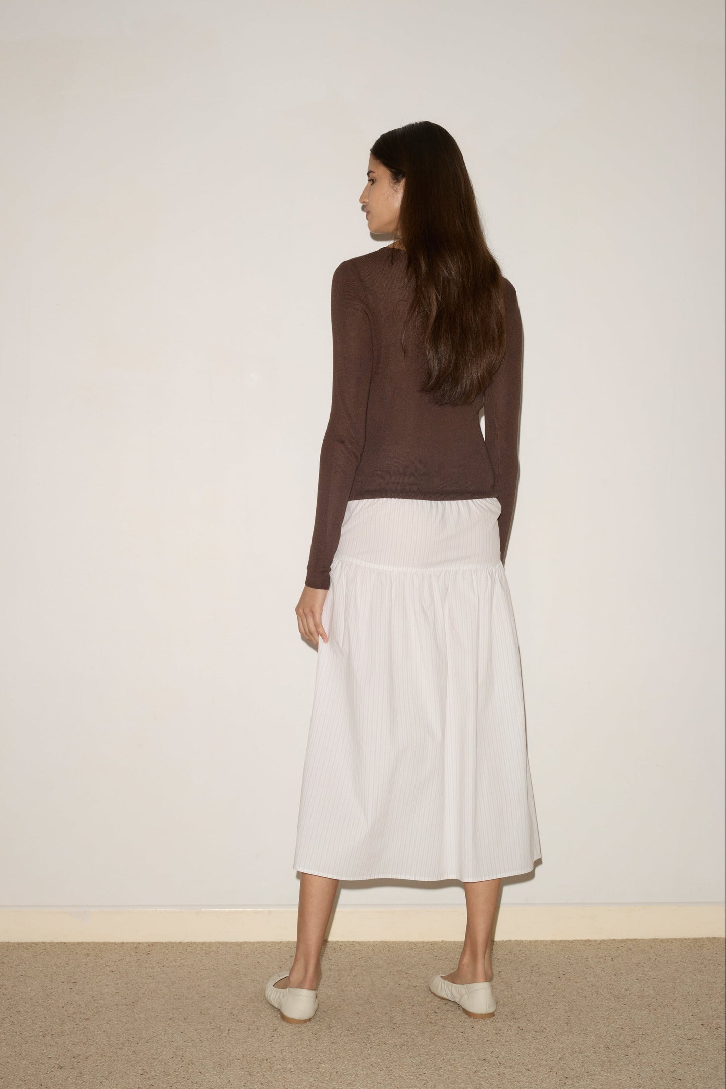 A woman with long brown hair stands facing a plain white wall, wearing a brown long-sleeve top, white shoes, and the Button Skirt - Pacific Stripe by 0. The skirt features an elasticated waistline and the scene is minimal indoors.