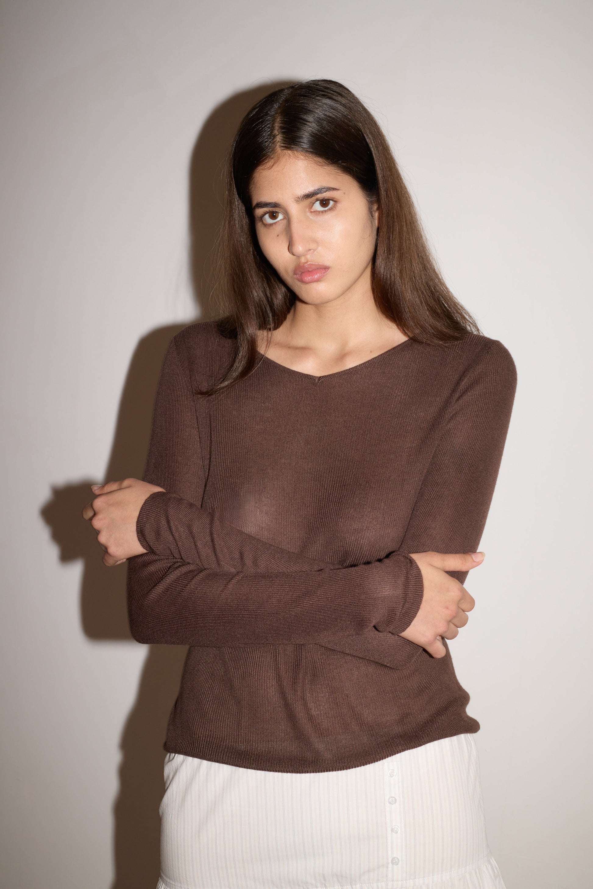 A young woman with long brown hair stands against a plain wall, wearing the 0 Knitted Top - Dark Brown and a white skirt. She folds her arms and gazes seriously at the camera as shadows fall on the wall behind her.