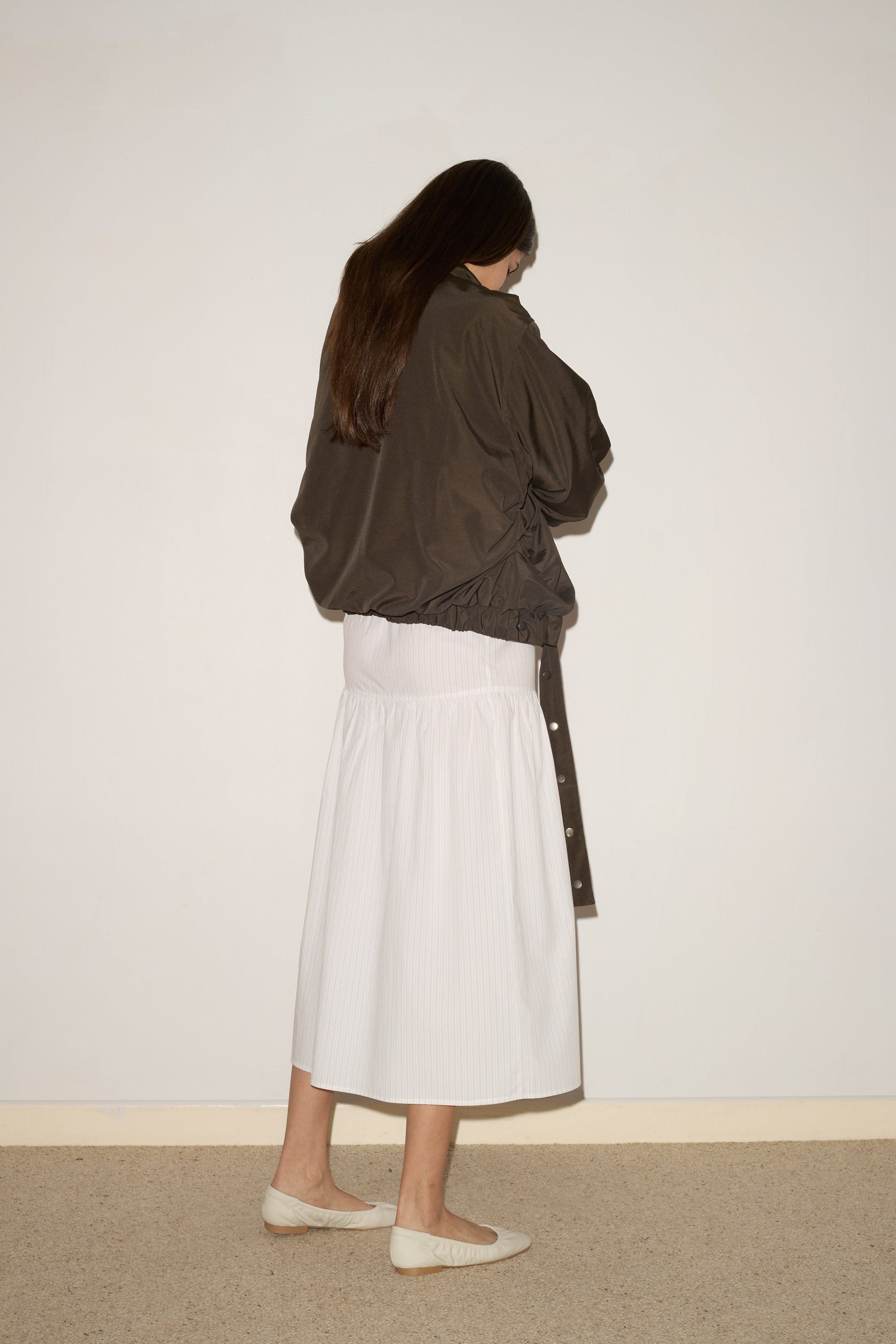 A person with long brown hair faces a wall, wearing the 0 Press Stud Jacket - Dark Chocolate, a long white skirt, and white flat shoes on a light carpeted floor against a plain off-white background.